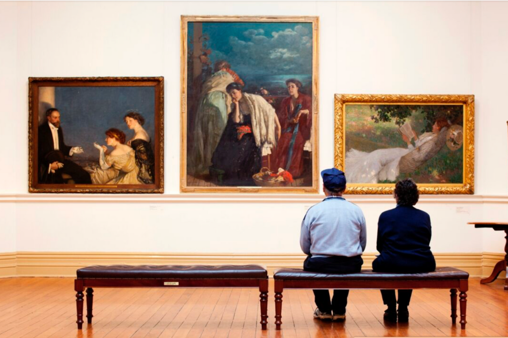 Two people sitting on a bench in an art gallery, viewing three large paintings on the wall.