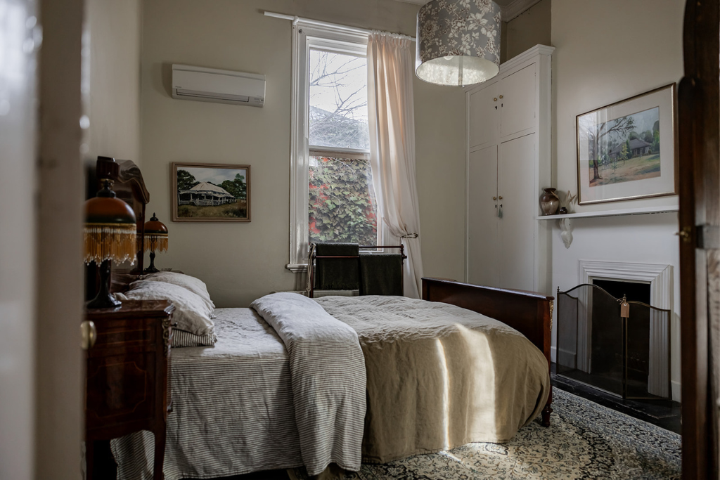 A cozy bedroom with a wooden bed, beige bedding, framed artwork on the walls, a window with white curtains, a fireplace, and decorative items on the mantle.