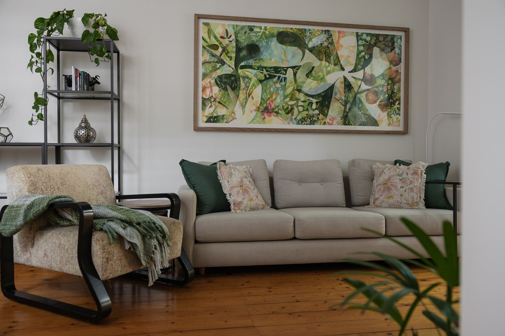 Living room with a beige sofa and pillows, a floral armchair with a blanket, a black metal bookshelf with decorative items and books, a large abstract botanical painting on the wall, with hardwood floors and potted plants.