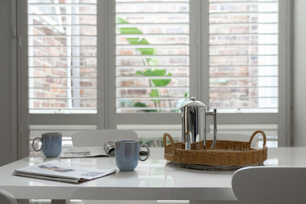 A modern dining table with two blue coffee mugs, an open newspaper, a silver coffee pot on a wicker tray, and a magazine, set in front of a window with white blinds and a view of a brick wall and a green plant.