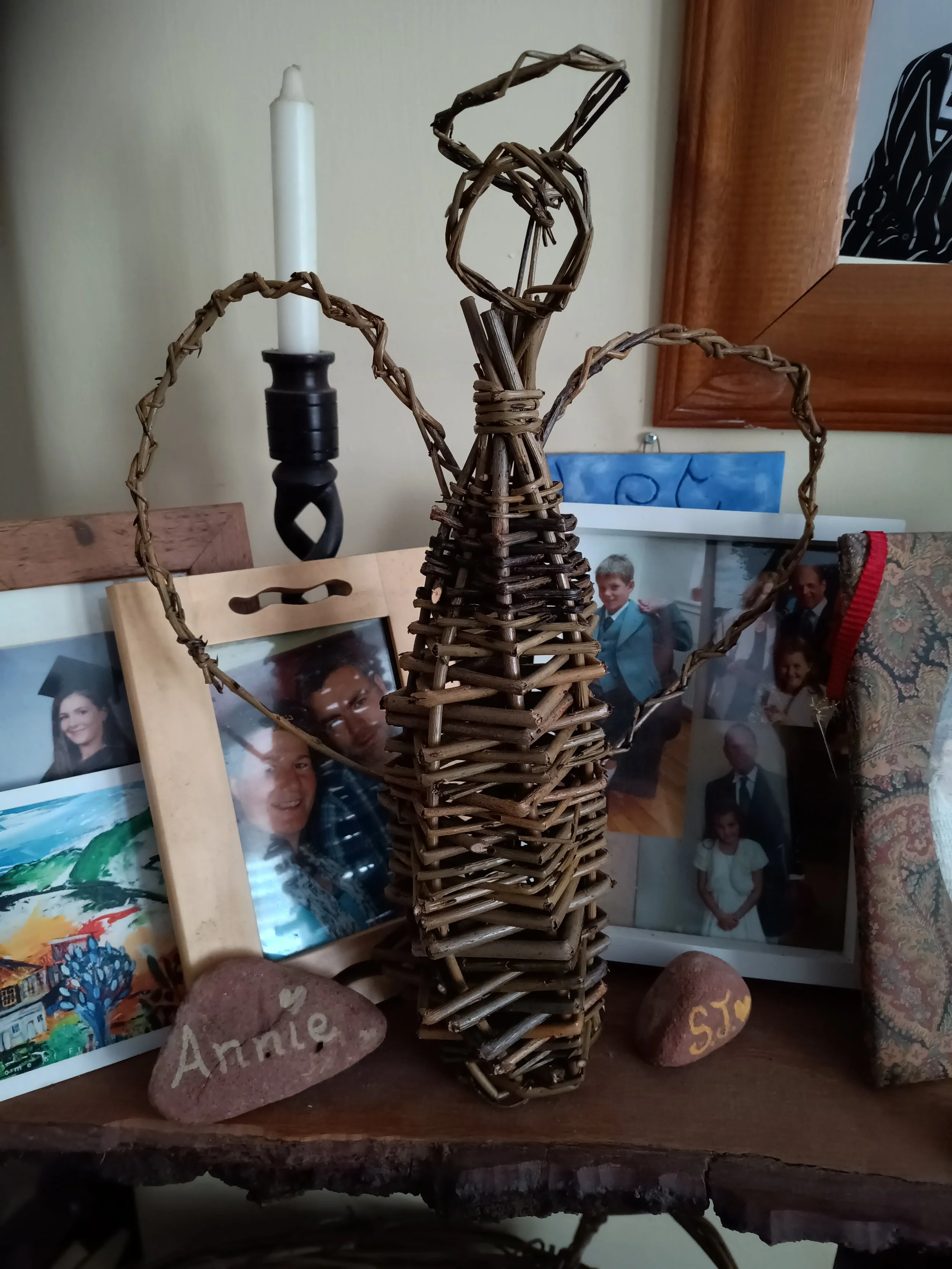Decorative woven willow wicker angel figurine by awaywithwillow.com on a wooden tabletop with framed family photos and painted stones, in a cozy home setting.