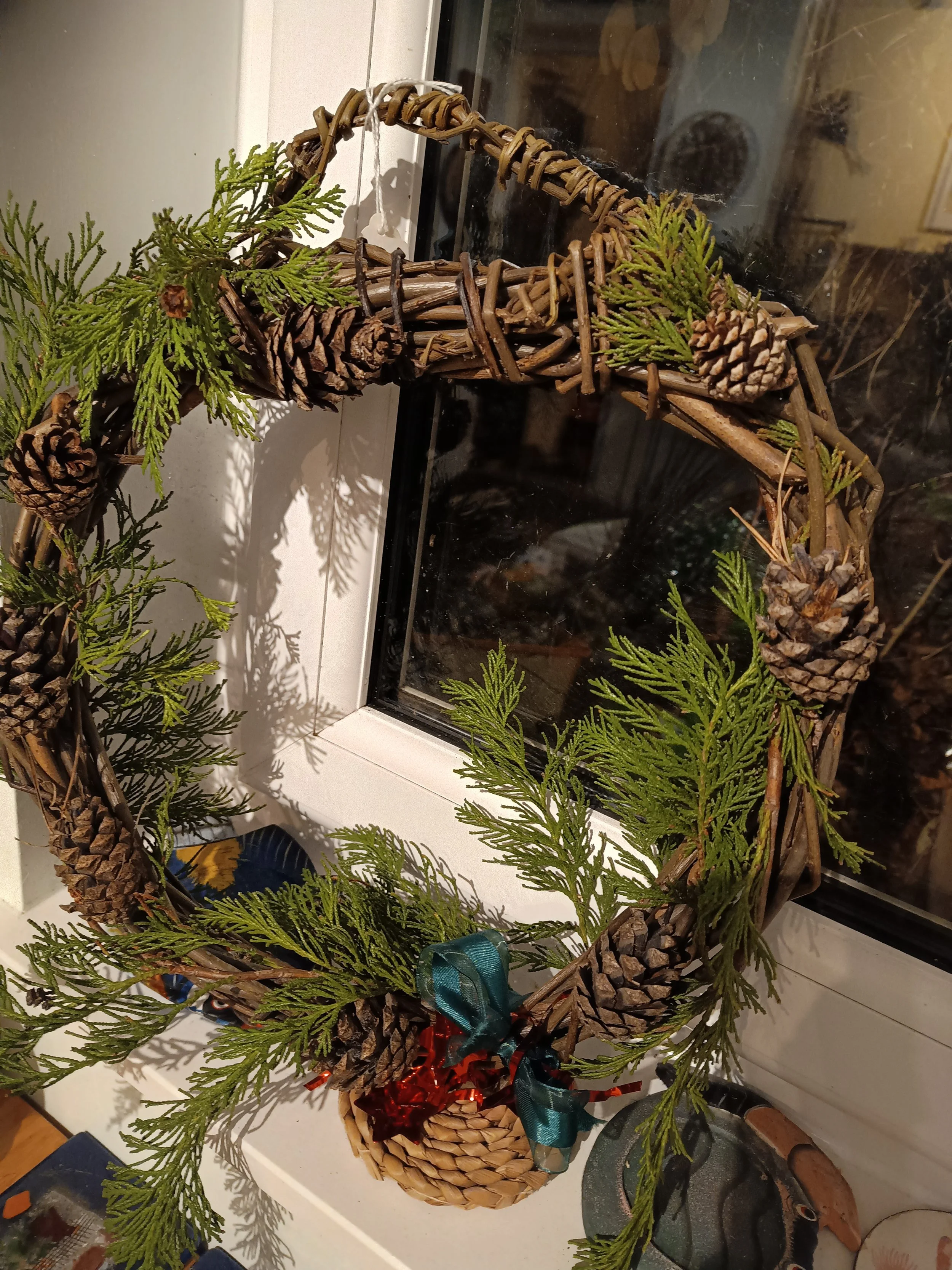 A Christmas willow based wreath decorated with pine cones, green foliage, and a small basket with a ribbon, hanging on a window.