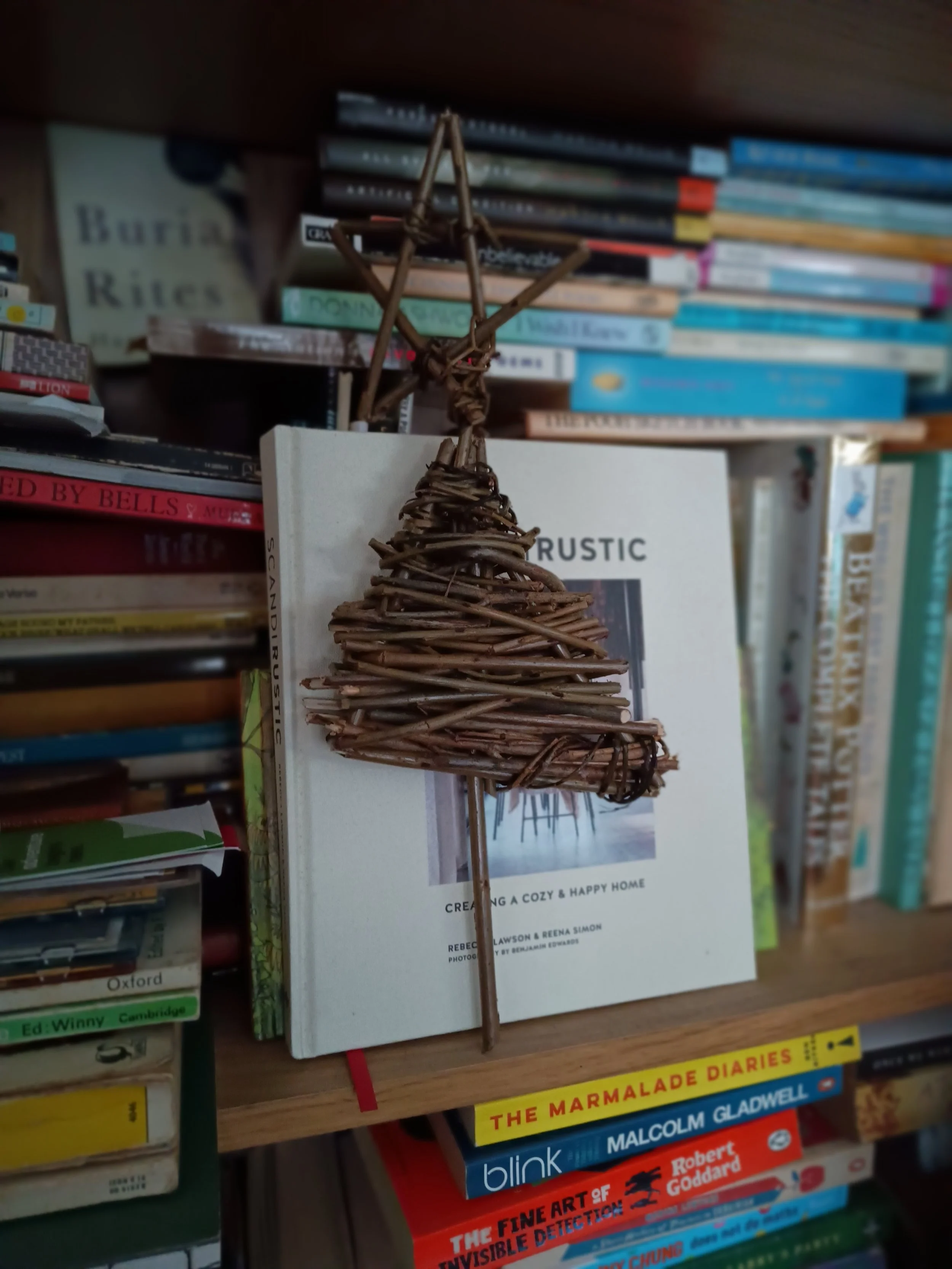 A woven willow wicker Xmas tree decoration with a star on top of the tree by awaywithwillo.com in front of a book titled 'Rustic: Creating a Cozy & Happy Home' surrounded by other books on a wooden shelf.