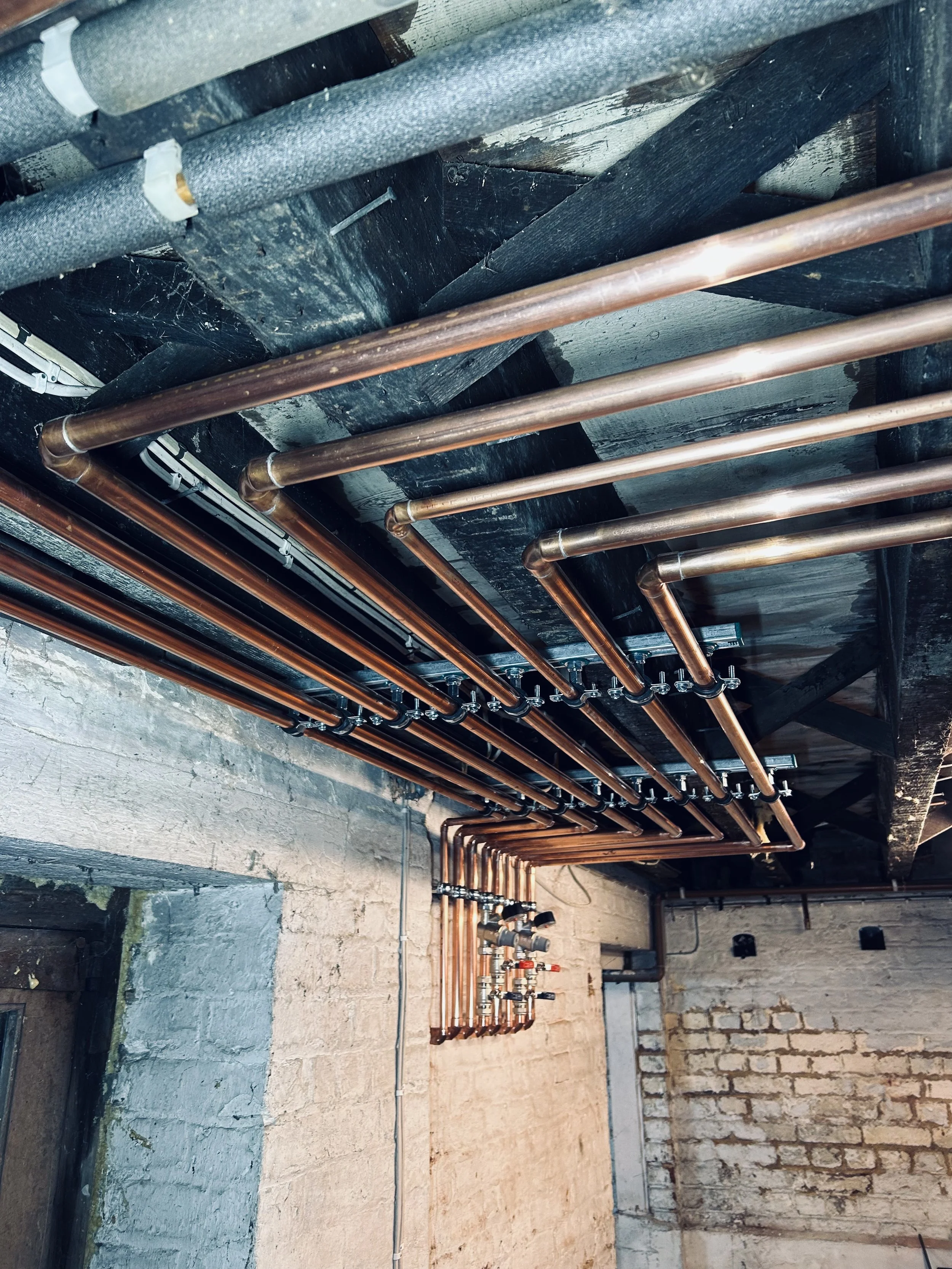 Copper pipes and fittings installed on a ceiling with black support beams and brick walls.