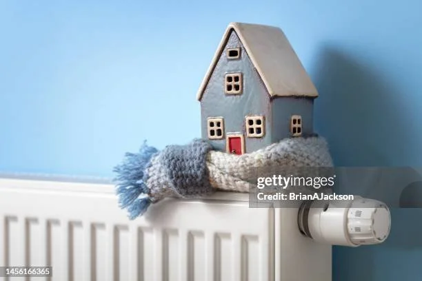 Small ceramic house placed on a radiator with a gray cozy scarf in front of a blue wall.
