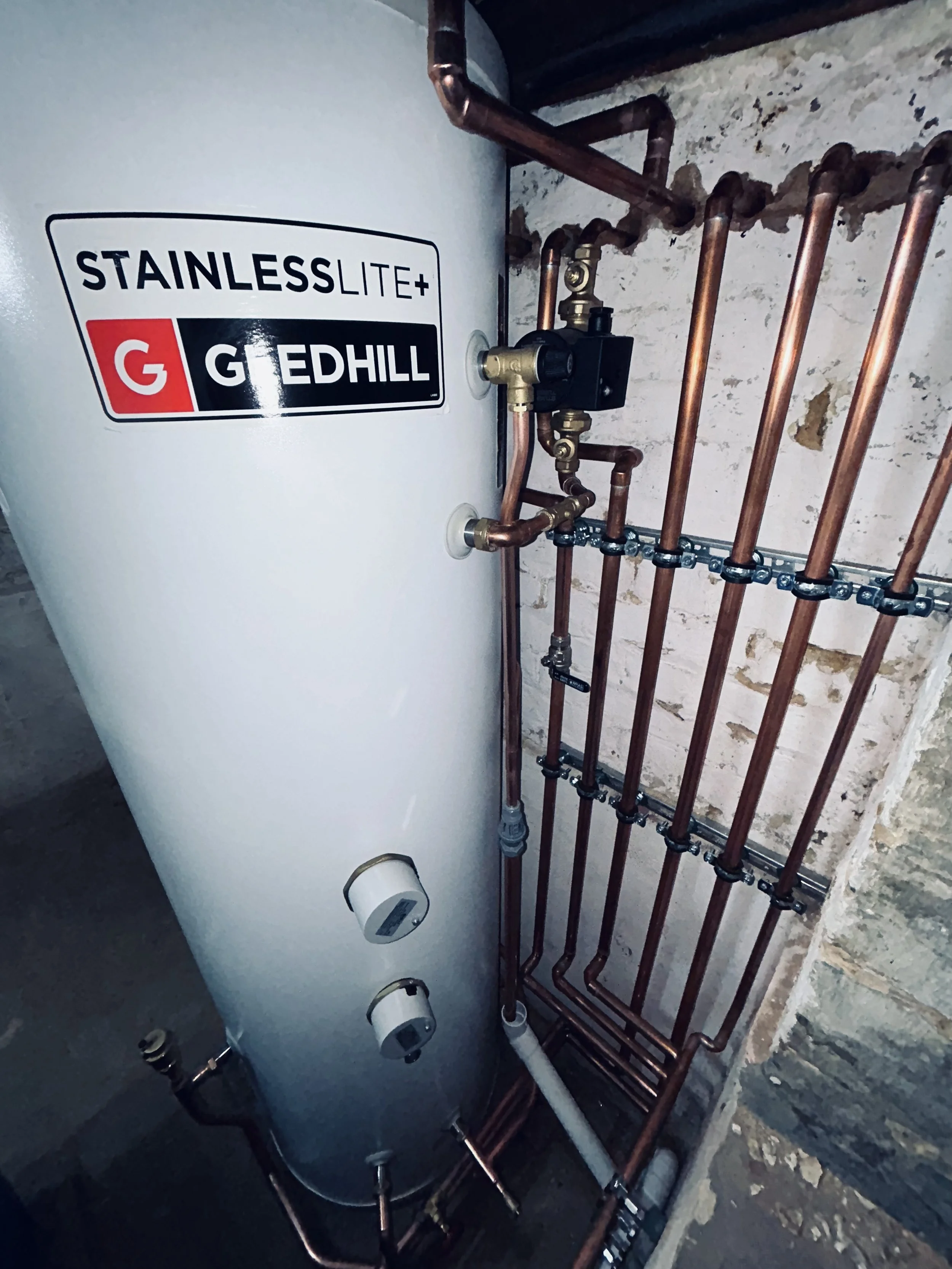 A water heater with copper pipes connected to it, mounted on a wall. The water heater has a label that reads 'Stainless Lite+ G Edhill'.