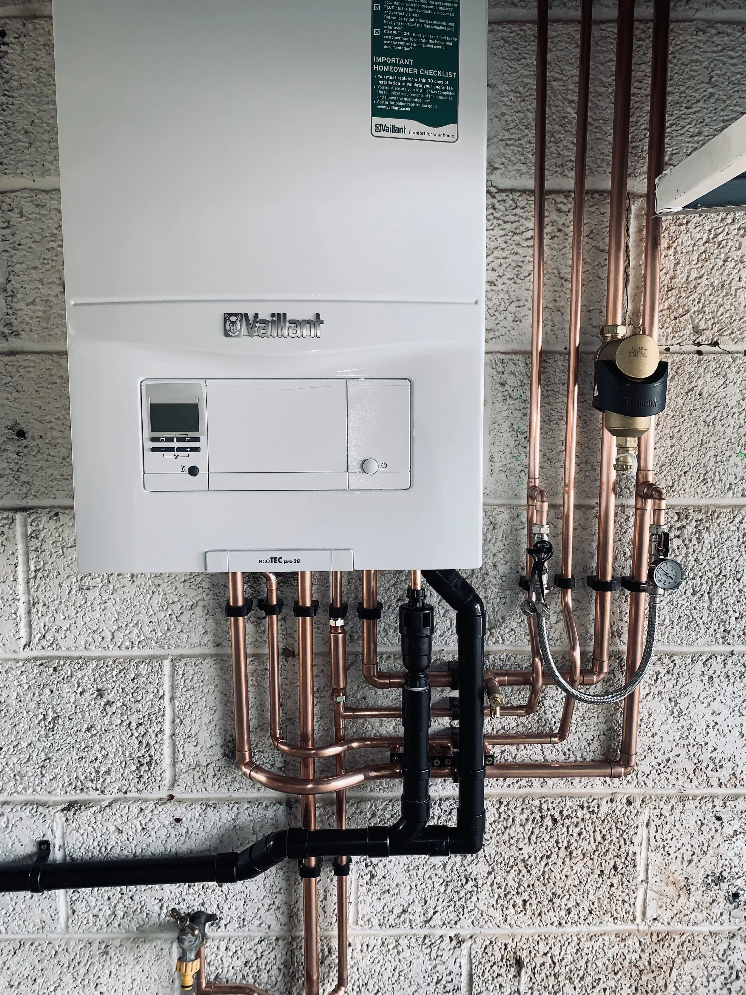 A white Vaillant boiler mounted on a cinder block wall with copper and black piping underneath, including a pressure gauge and shut-off valves.