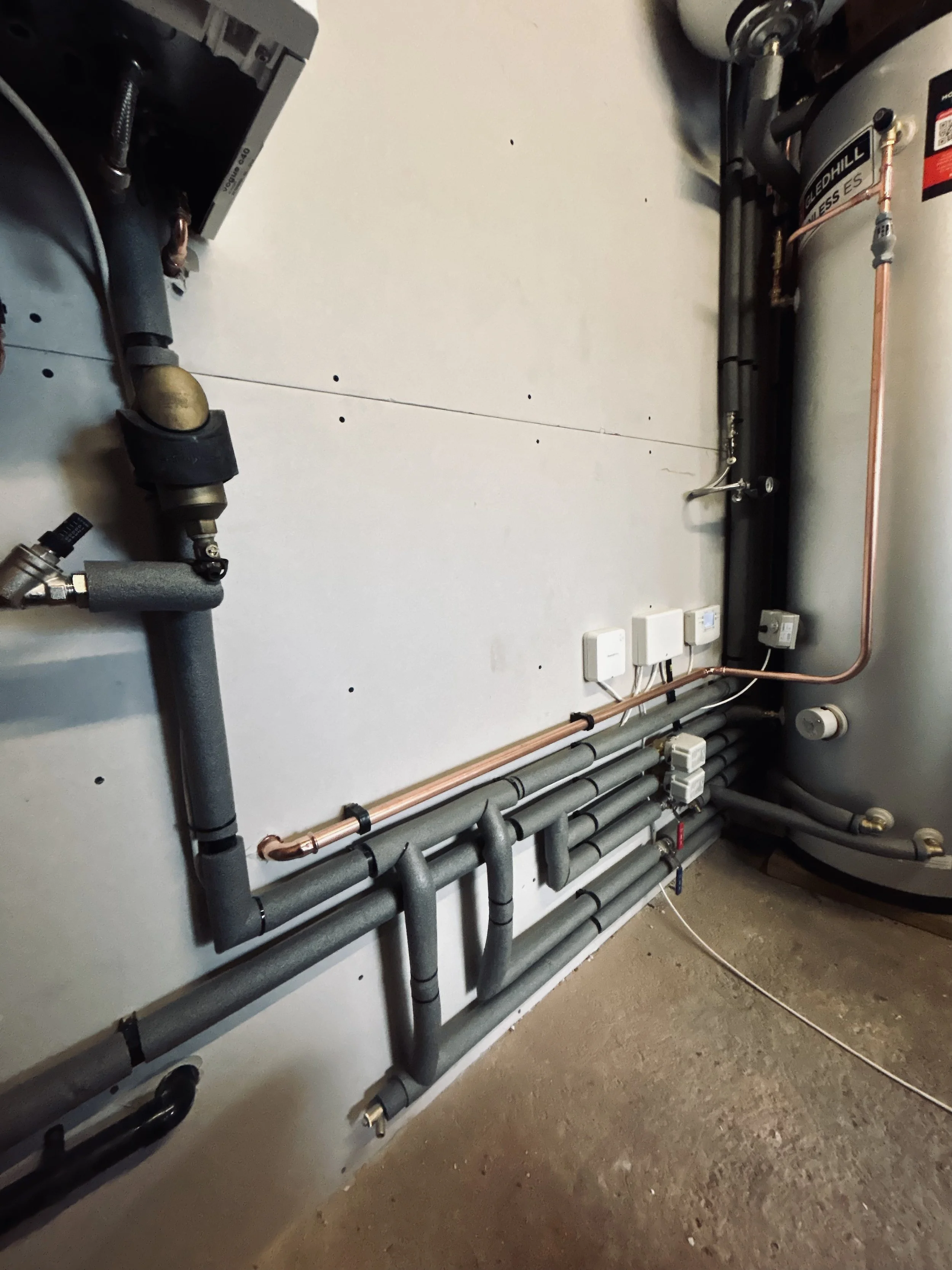 A utility room with various pipes, electrical outlets, and a large water heater tank.