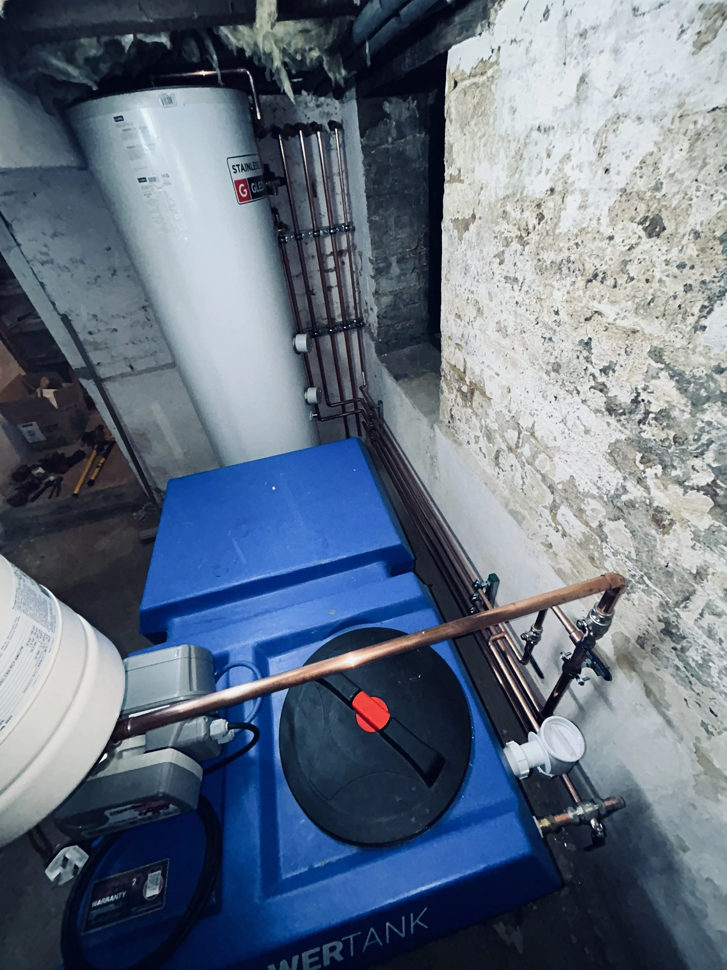 A water heater next to copper piping and a blue water storage tank in a basement with exposed brick walls.