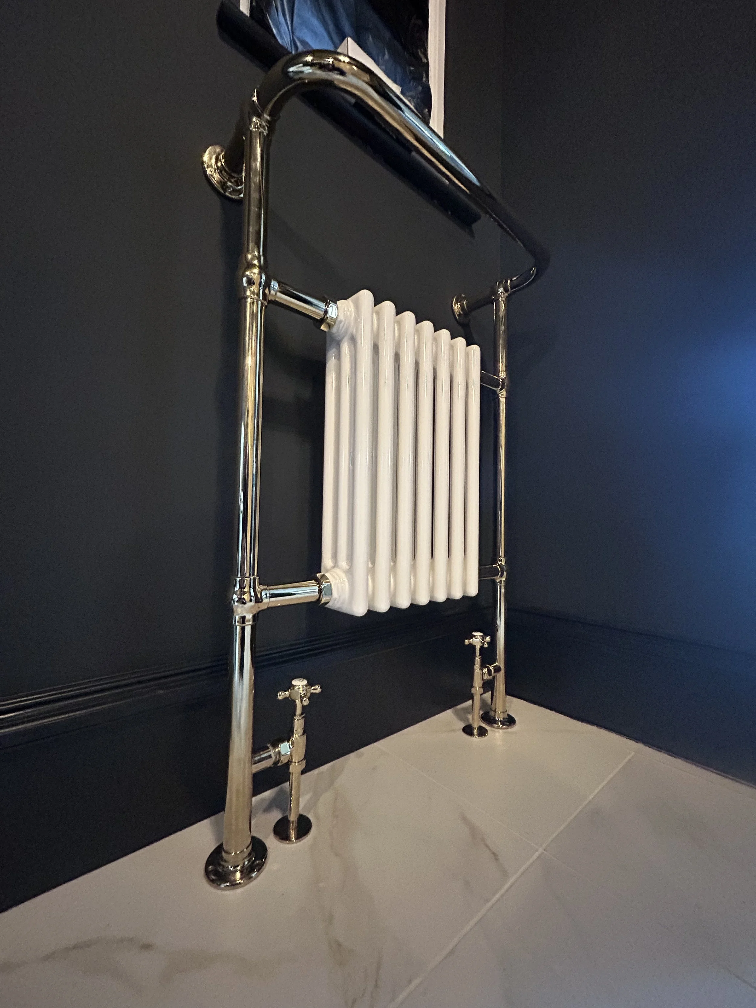 A modern radiator towel warmer with chrome finish mounted on a dark wall in a tiled bathroom.