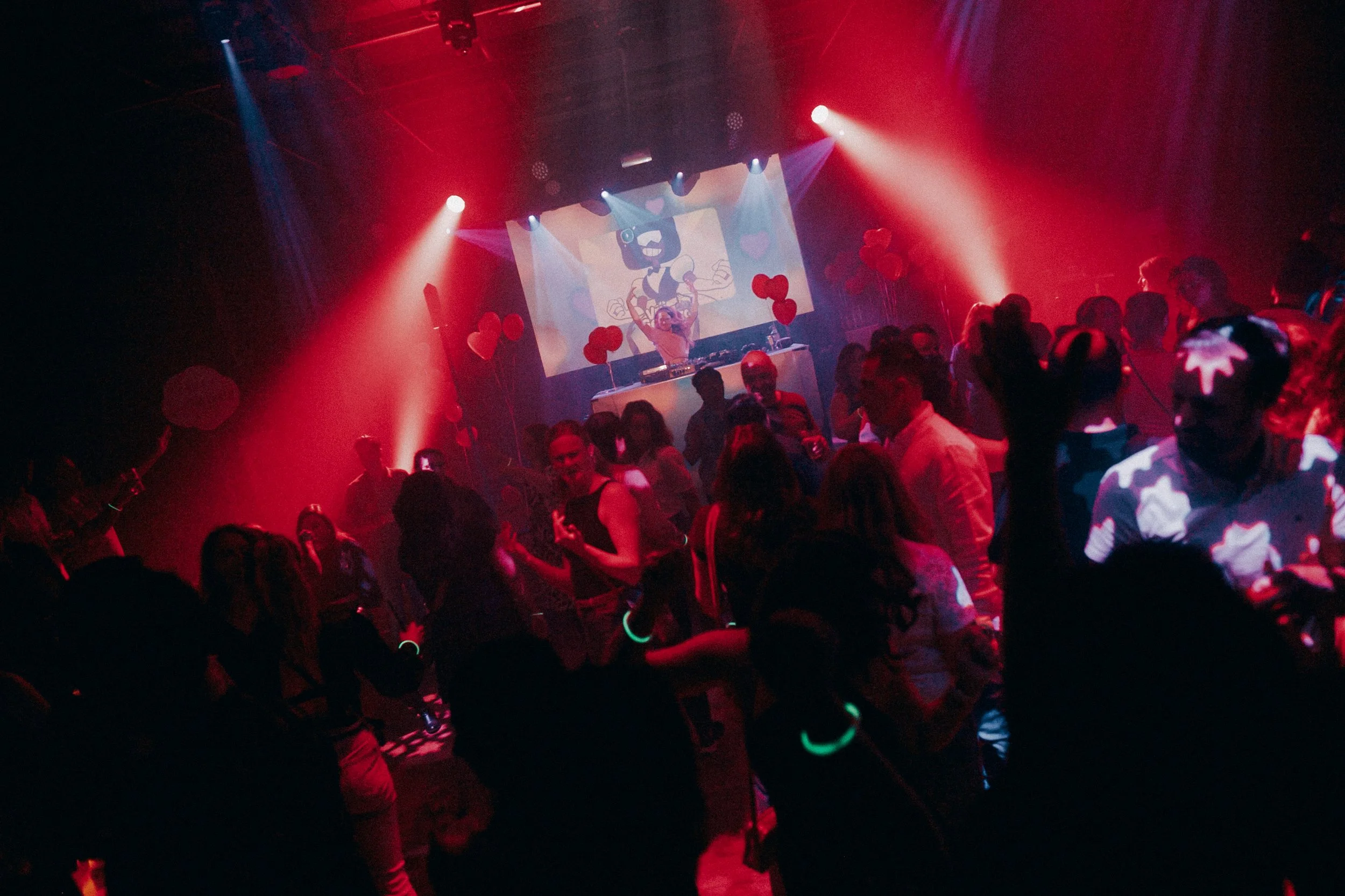 Crowd of people dancing at a nightclub with red lighting, a DJ on stage, and a screen displaying animated graphics in the background.
