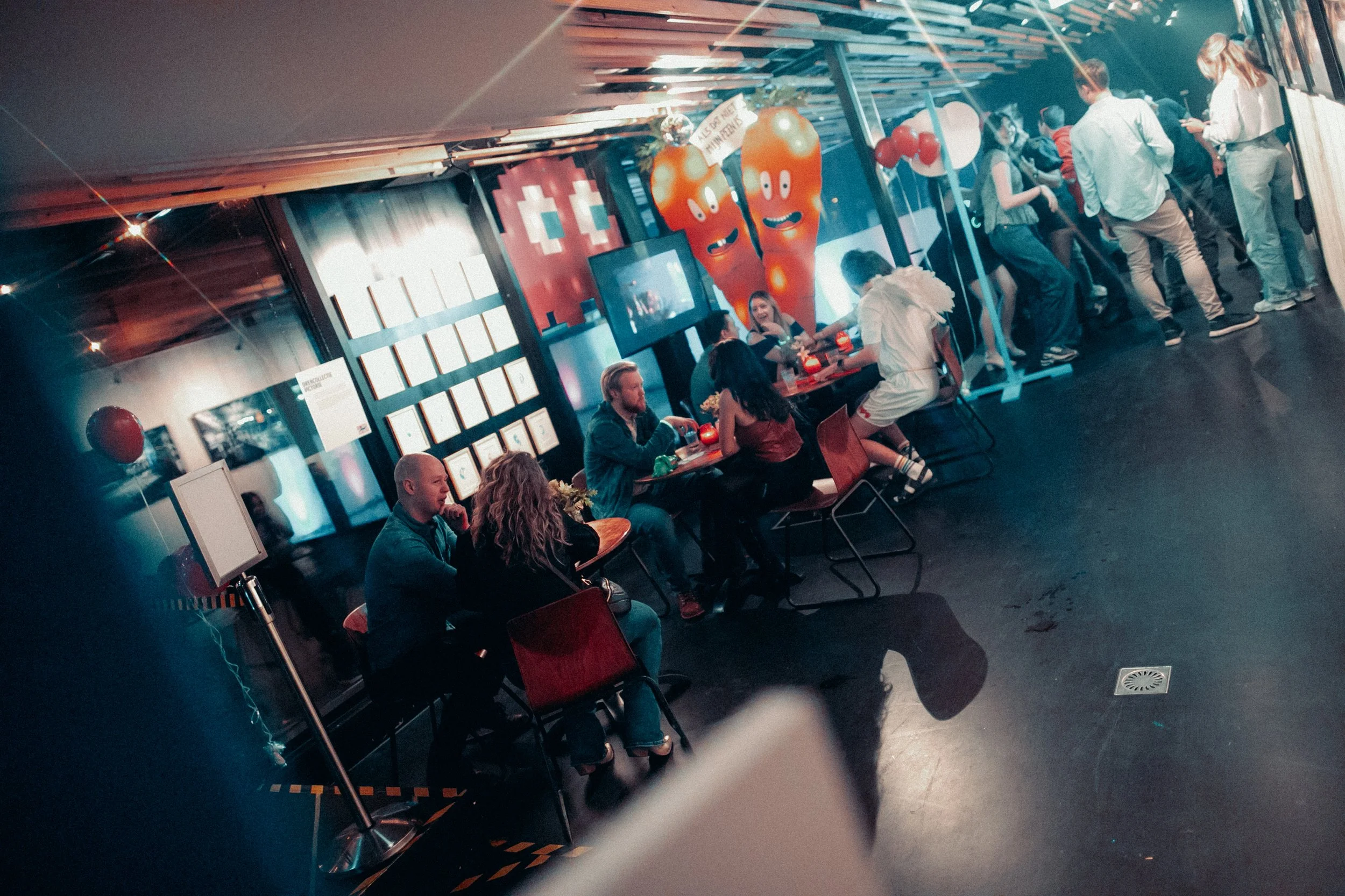 People sitting at a table, chatting, with a wall decorated with cartoon vegetables and balloons in the background.