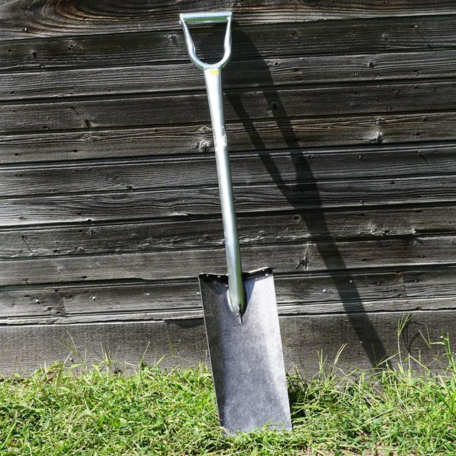A shovel with a metal blade and handle, planted in the ground in front of a wooden fence, with grass at its base.