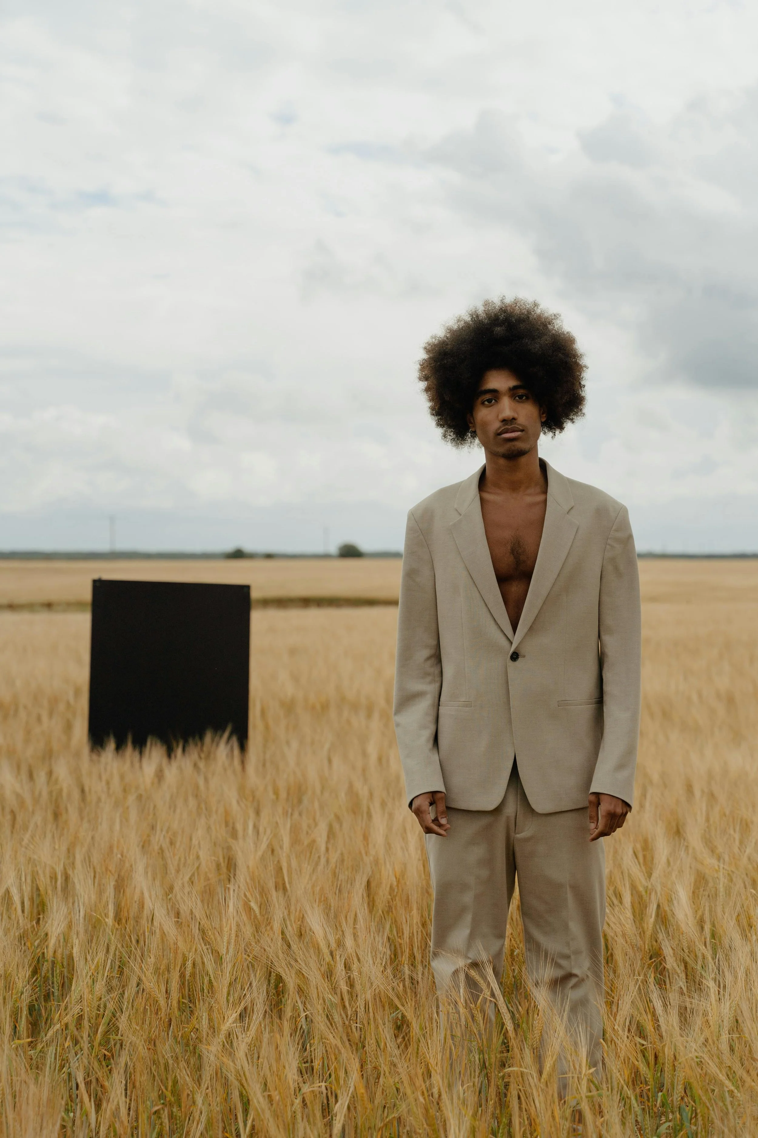 A young man with an afro hairstyle stands in a wheat field wearing a beige suit jacket and matching pants with no shirt underneath. There is a black rectangular object in the background and cloudy sky overhead.