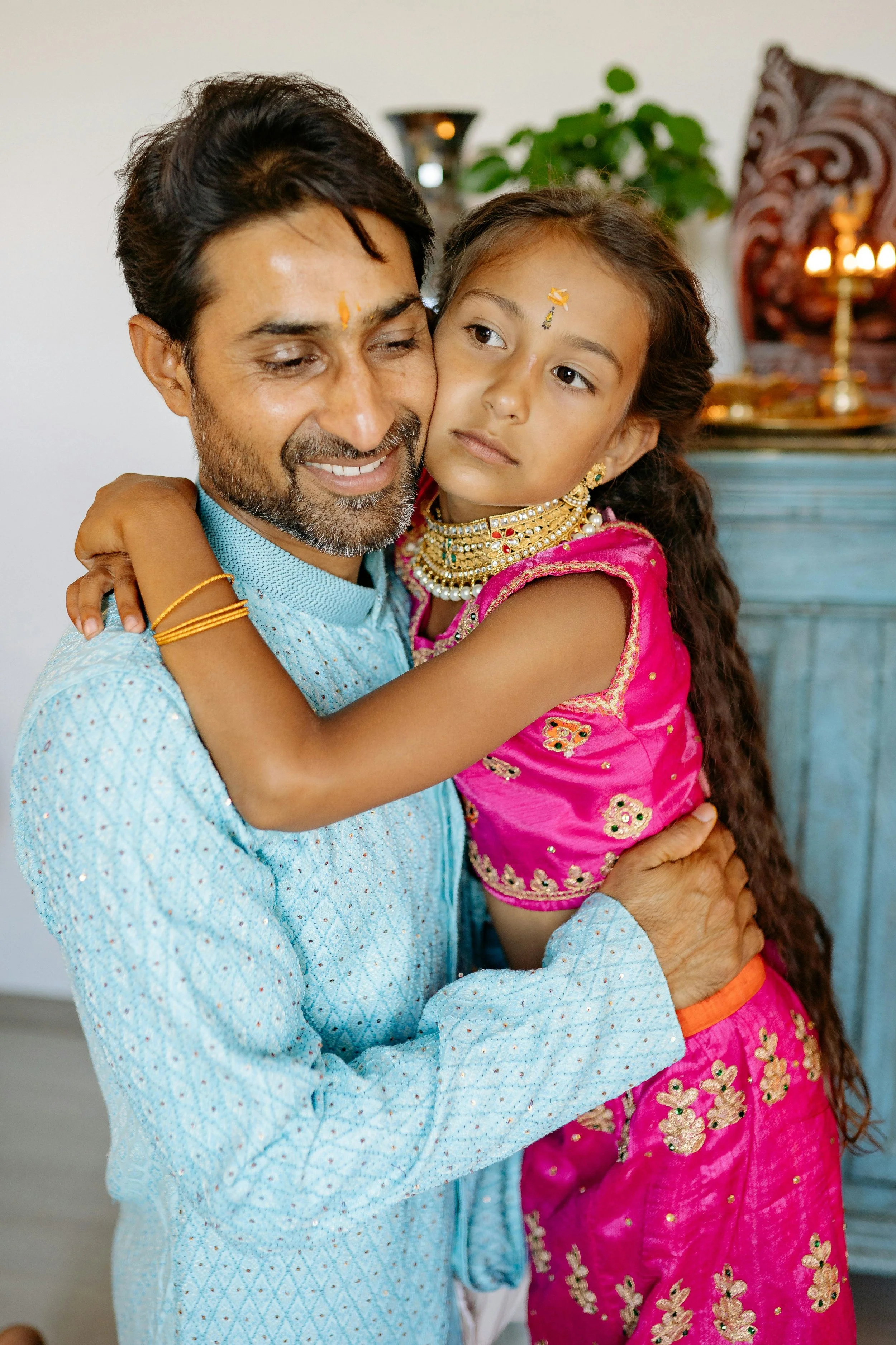 A man and a girl hugging each other, dressed in traditional Indian clothing, celebrating a cultural or religious event with decorative jewelry and a tikka on their foreheads.