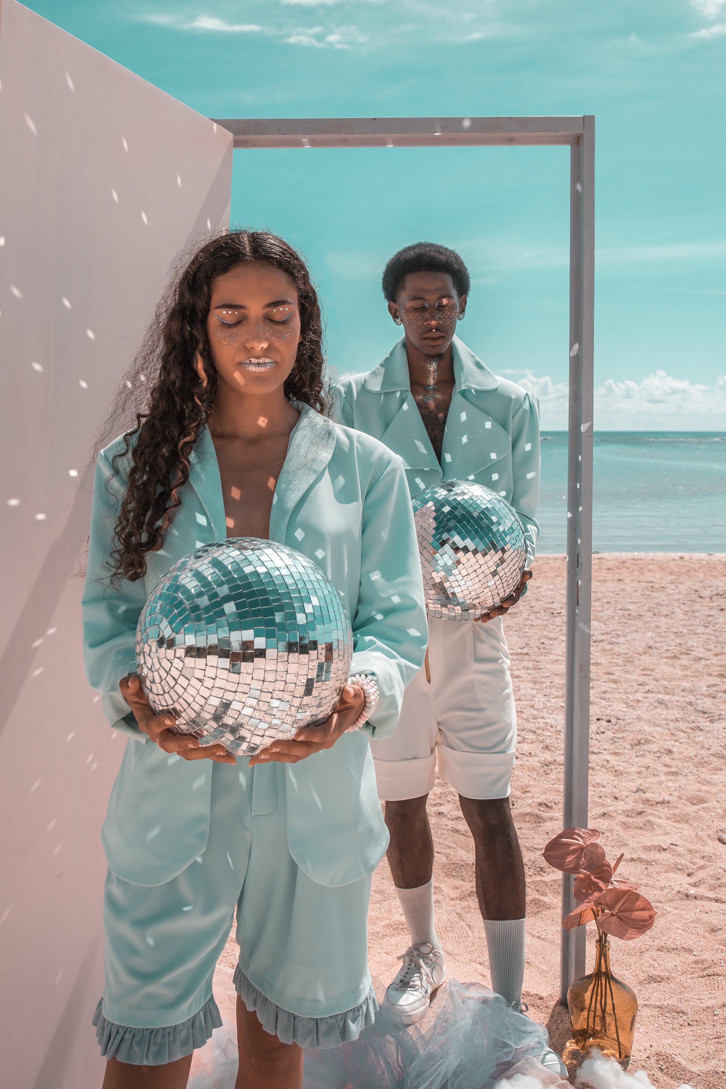Deux personnes debout sur une plage, tenant des boules disco réfléchissantes, sous un décor minimaliste avec un vase en verre et une plante. Le ciel est bleu avec quelques nuages.