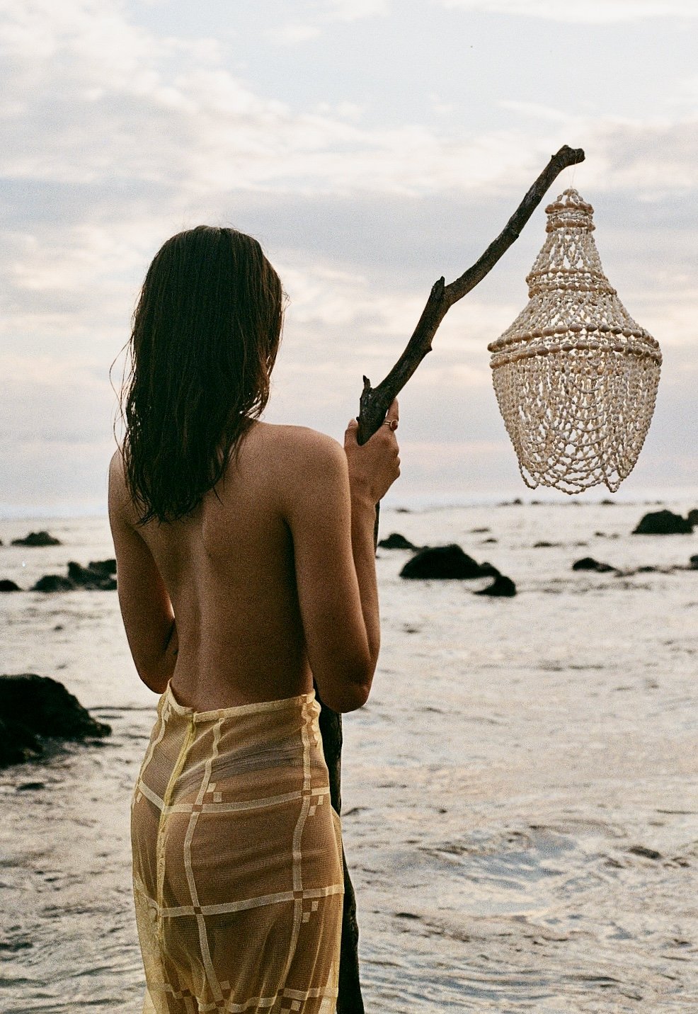 Une femme nue vue de dos, tenant un bâton avec une lampe de chandelier en décoration suspendue, sur une plage rocheuse au coucher du soleil.