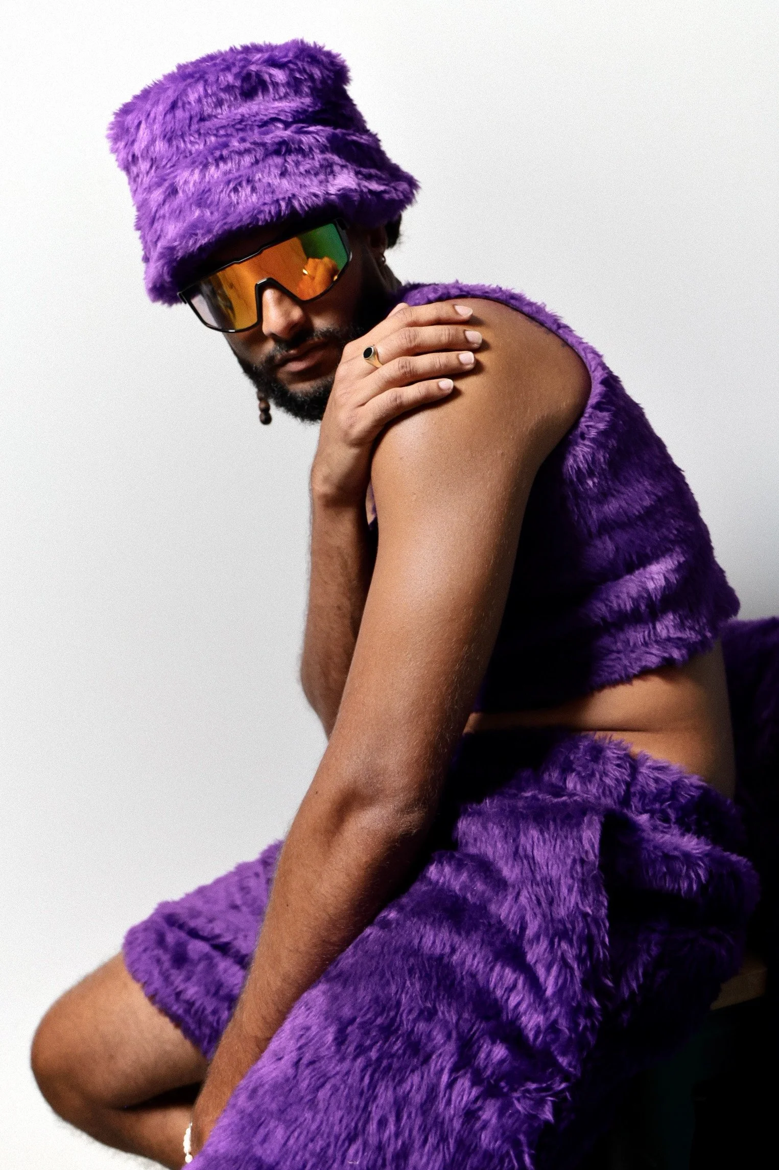 Homme portant un chapeau et un haut en fausse fourrure violette, avec des lunettes de soleil réfléchissantes, posé de manière stylée.