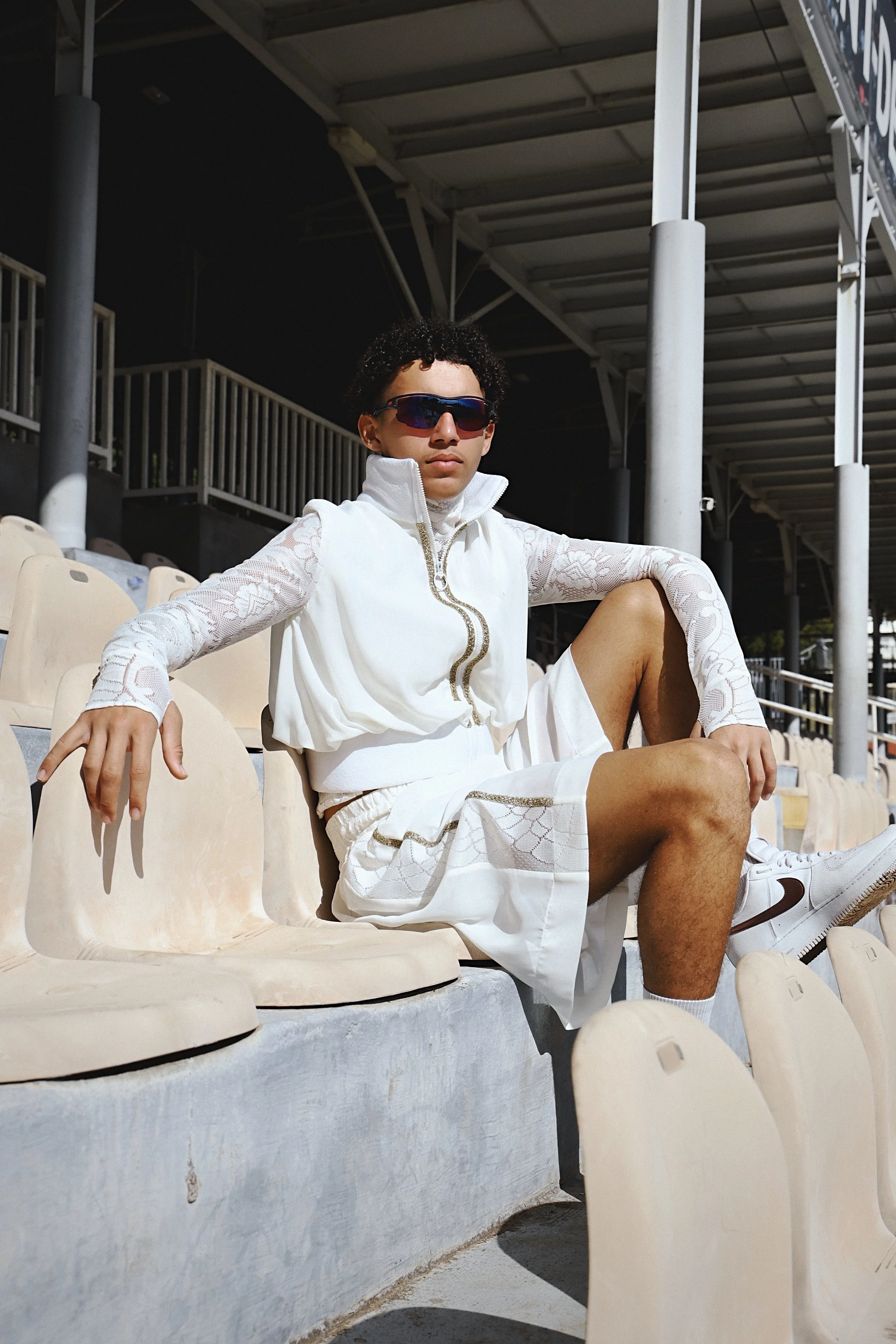 Un jeune homme assis dans les gradins d'un stade, portant des vêtements de sport blancs, des lunettes de soleil et un casque de soleil.