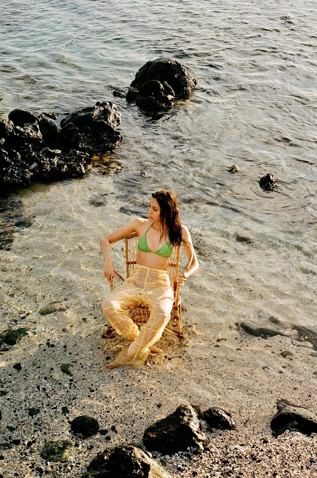 Une femme assise dans une chaise en rotin sur une plage, portant un haut de bikini vert et un pantalon léger beige, avec la mer en arrière-plan et des rochers dans l'eau.