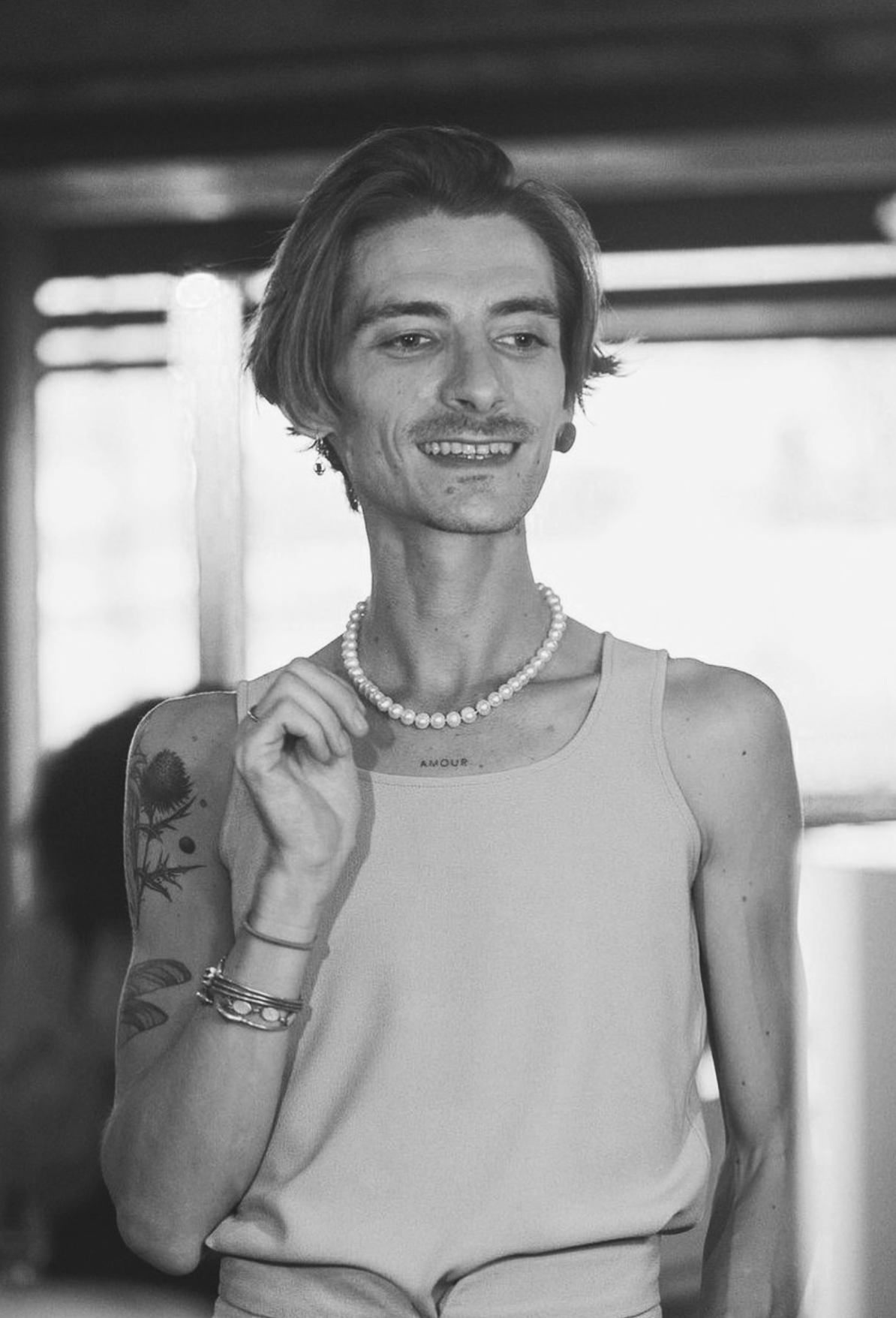 Une femme souriante portant un collier de perles, un tatouage sur le bras, un tank top, avec un fond flou et une lumière naturelle venant d'une fenêtre.