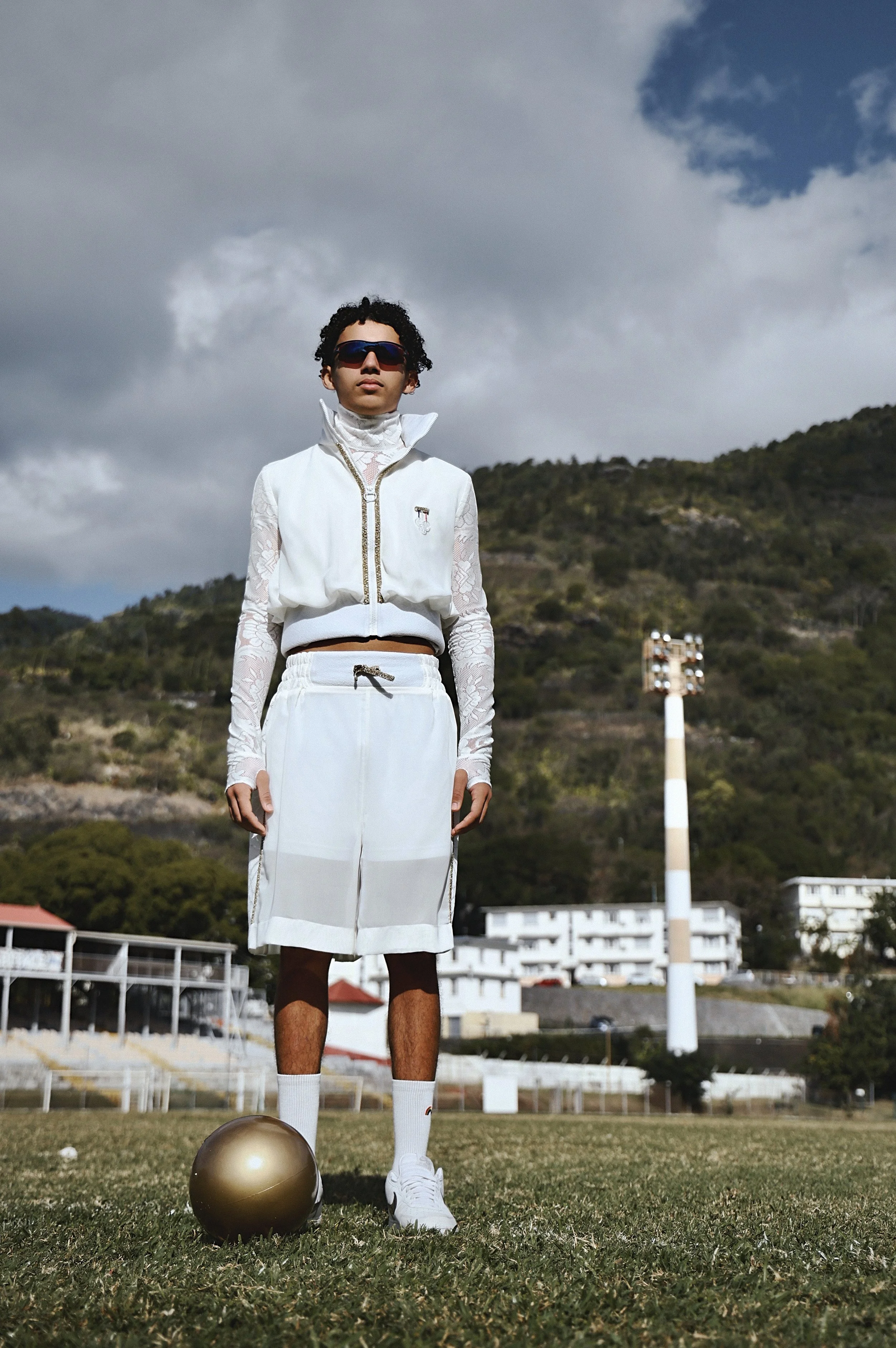 Jeune homme portant des vêtements de sport blancs, debout sur un terrain de football avec un ballon doré, montagne en arrière-plan, ciel nuageux.