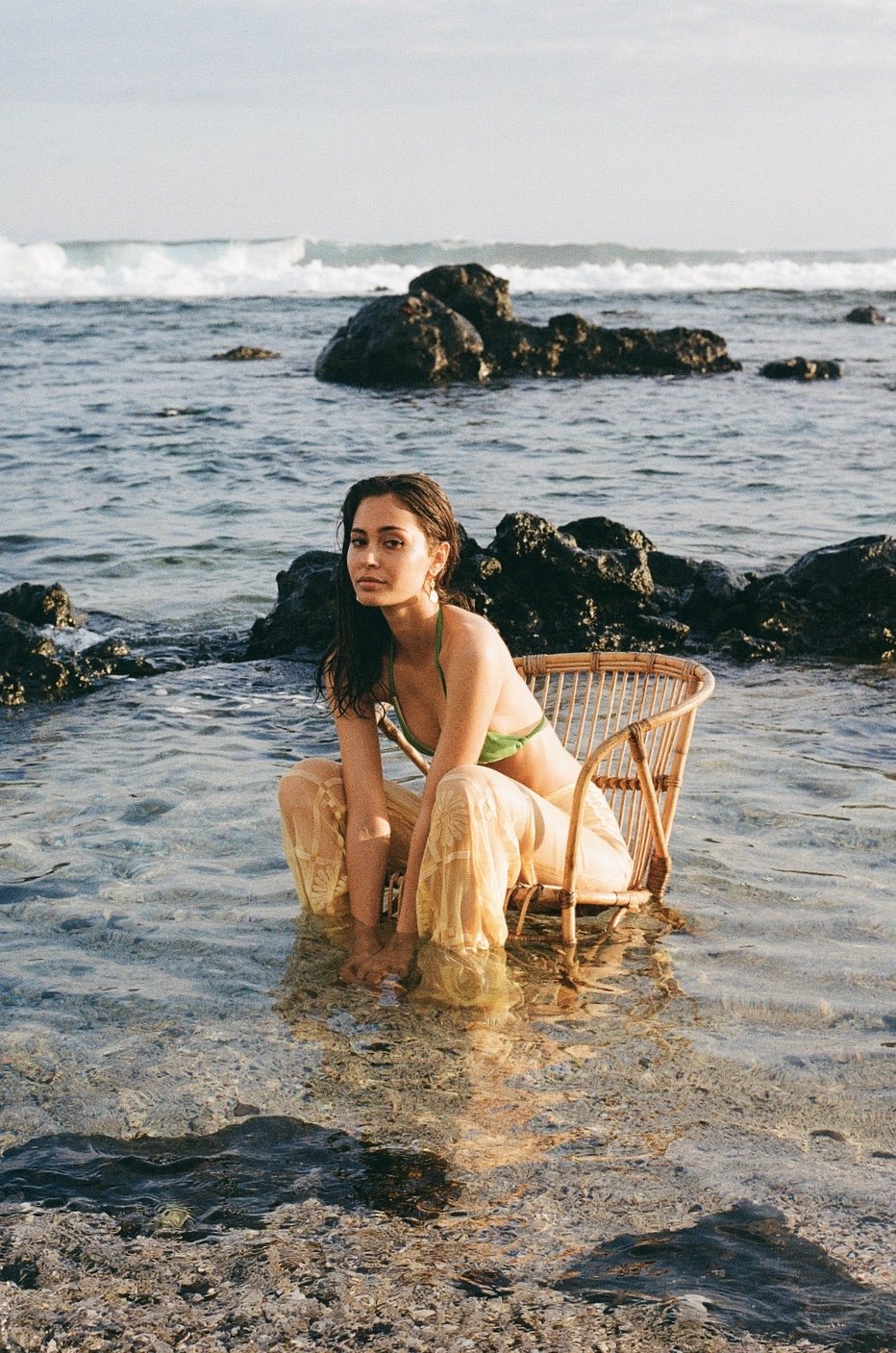 Jeune femme en maillot vert assise dans un fauteuil en rotin au bord de la mer, avec des rochers et des vagues à l'arrière-plan.