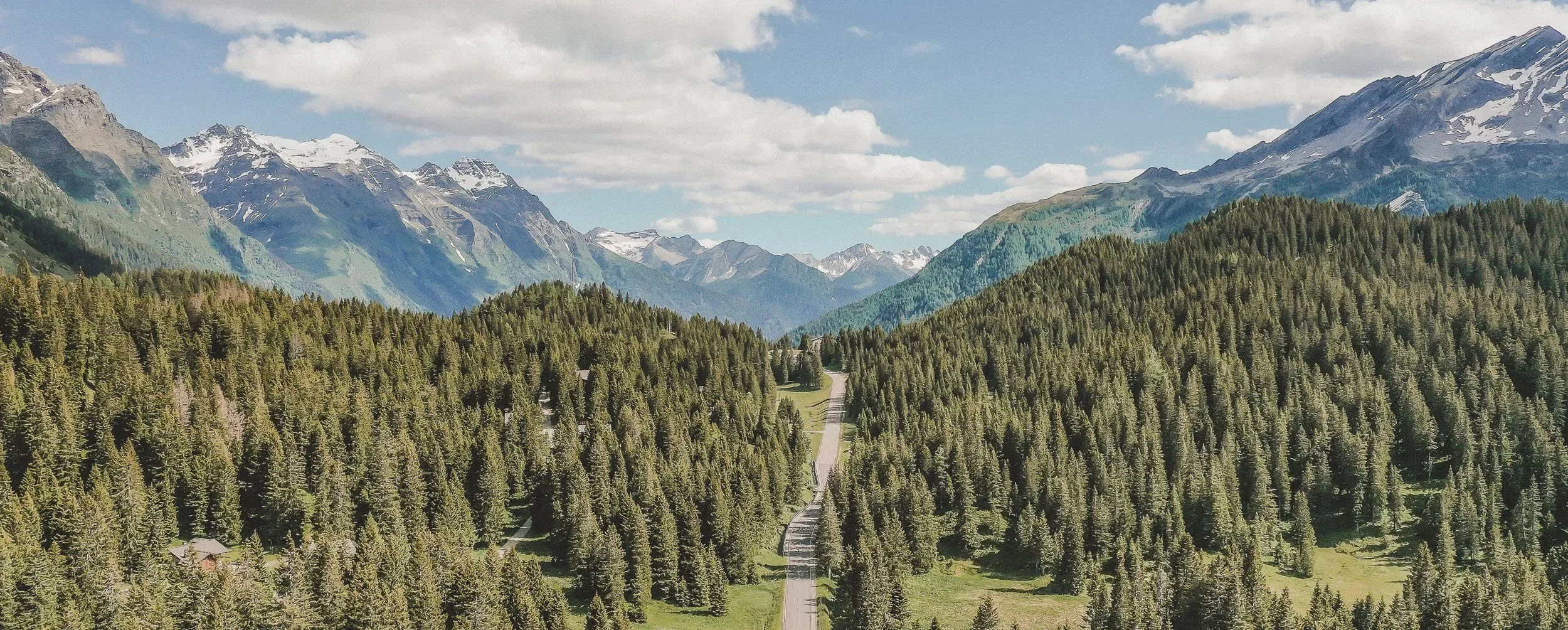 Veduta aerea estiva della valle di San Bernardino con fitti boschi e montagne alpine sullo sfondo.