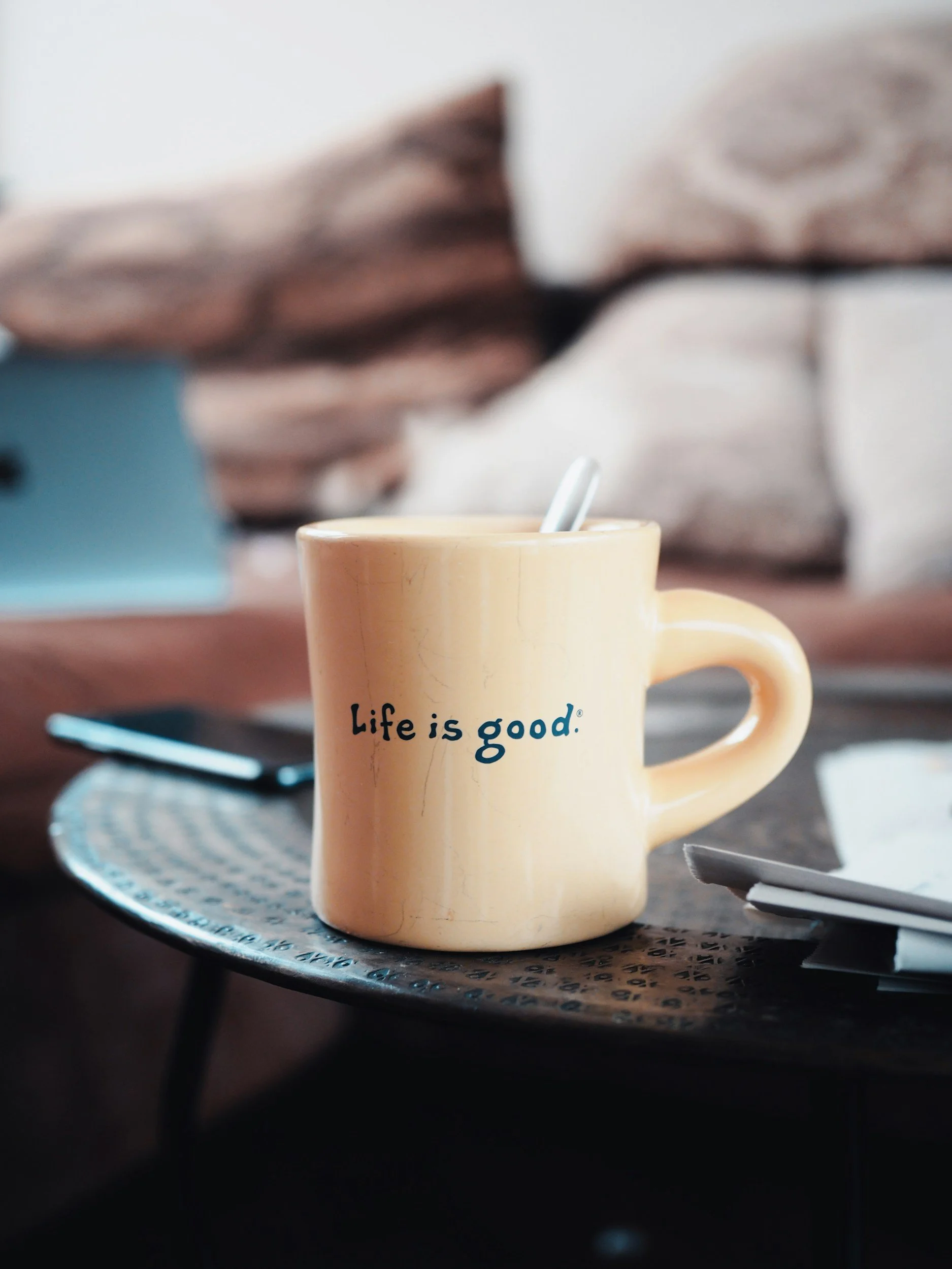 Yellow mug with black text reading 'Life is good' on a black patterned table holding a spoon. In the background, a smartphone and papers are visible with a blurred sofa and cushions.
