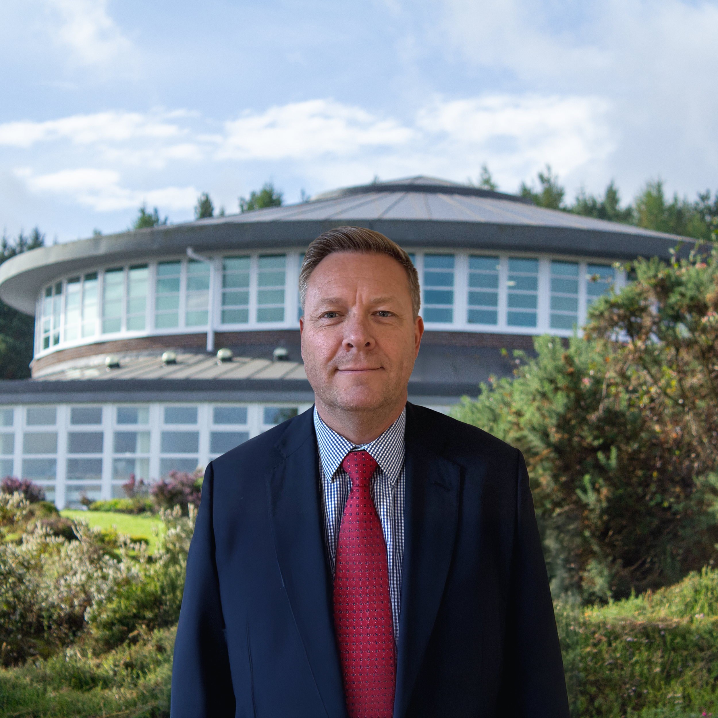 A man in a suit standing outdoors in front of a modern, circular building with large windows, surrounded by greenery and bushes, under a partly cloudy sky.