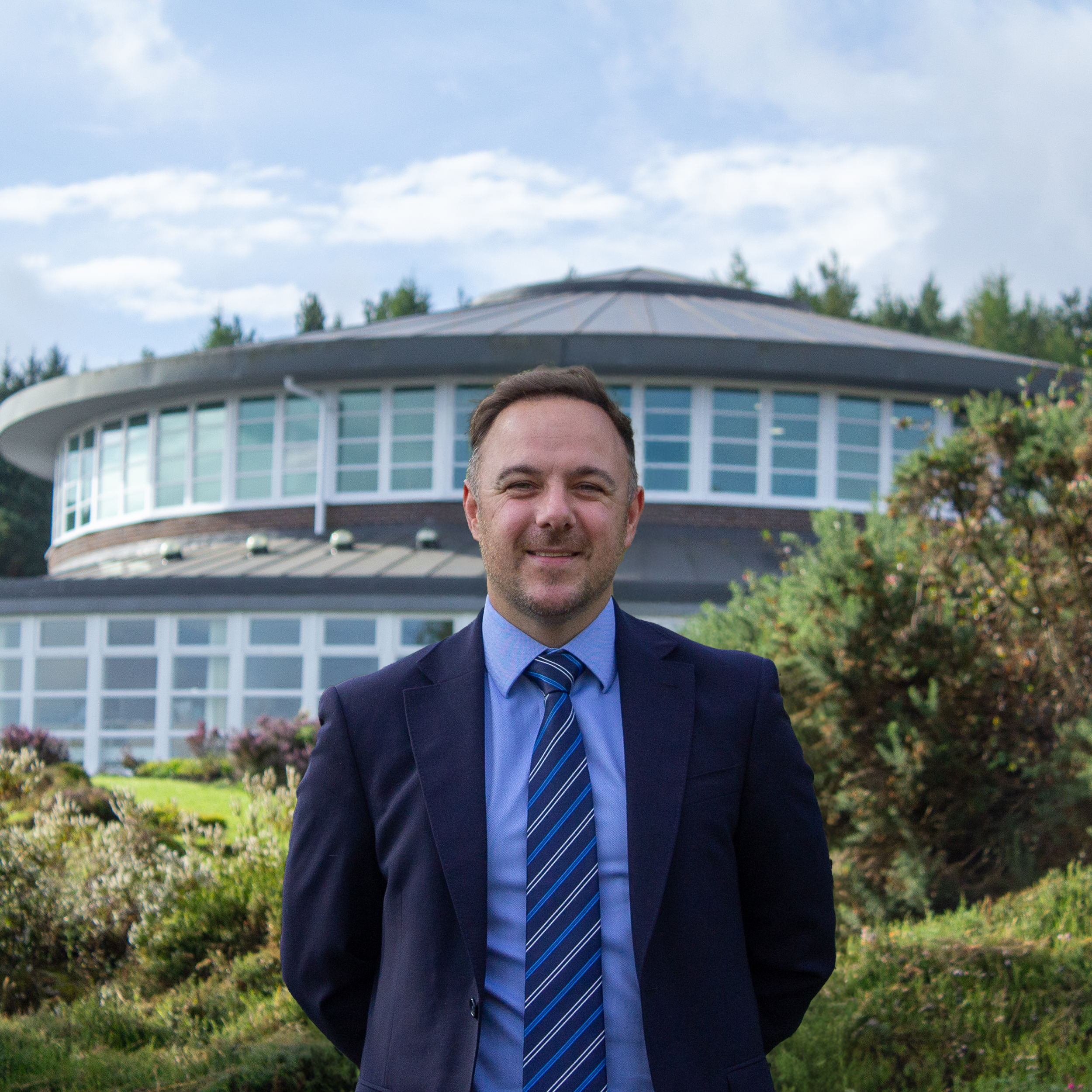A man in a navy suit and blue striped tie standing outside in front of a modern, circular building with large windows, surrounded by greenery.