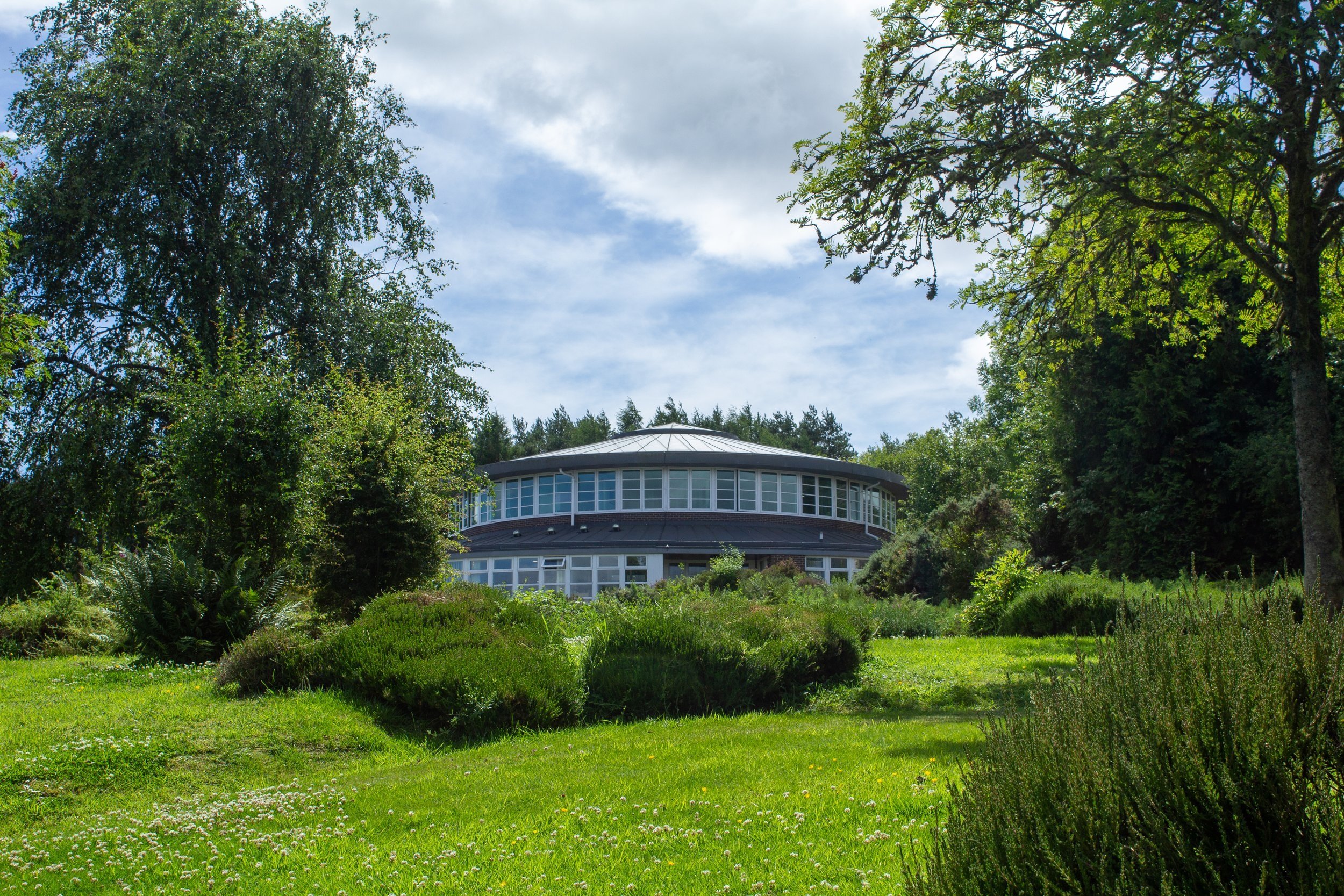 A round modern building with large windows is surrounded by lush green trees and a well-manicured garden on a sunny day with a partly cloudy sky.