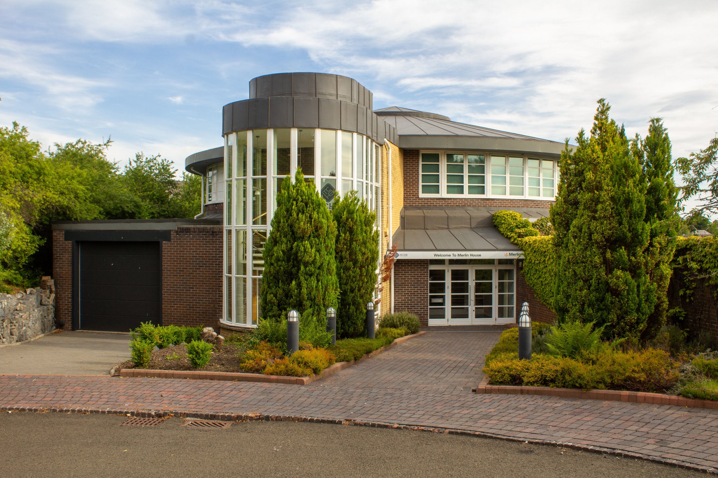 Modern multi-story brick building with large glass windows, surrounded by landscaped bushes and trees, with a paved driveway and sidewalk.