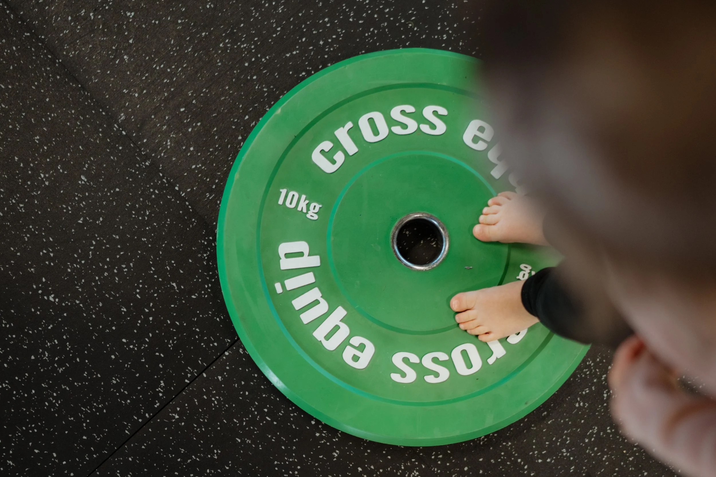 Fuß eines Kindes auf einer grünen CrossFIT-Kurzhantel mit 10 kg, auf schwarzem Boden