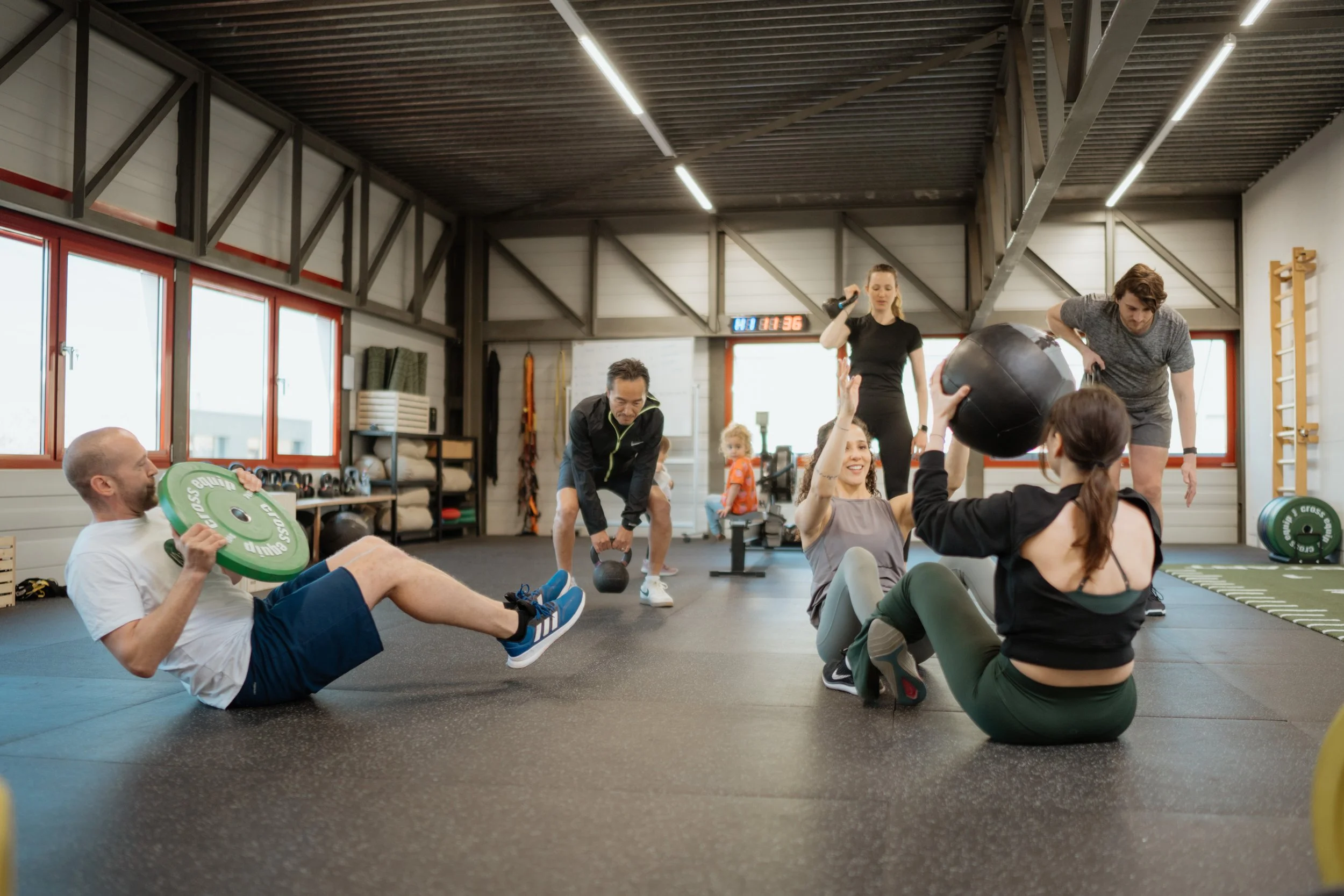 Menschen trainieren in einem Fitnessstudio, einige setzen sich auf den Boden mit Gewichten und Medizinbällen, während ein Trainer Übungen erklärt.