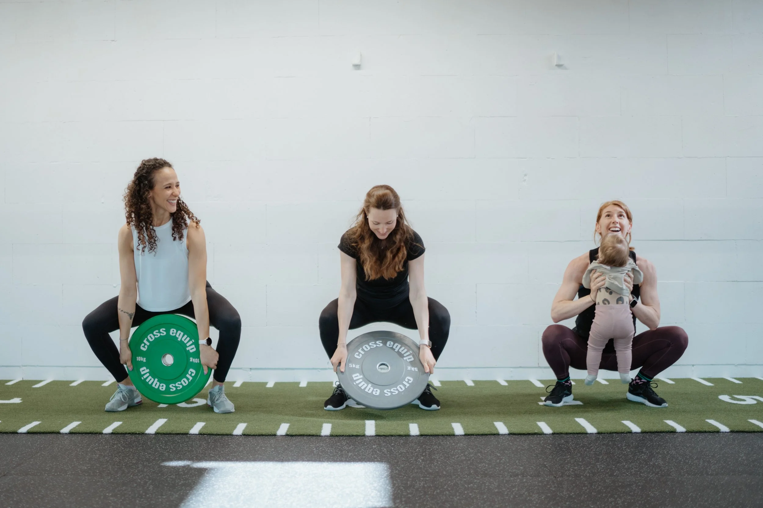 Drei Frauen beim Gewichtheben im Fitnessstudio, eine Frau hält ein Baby.