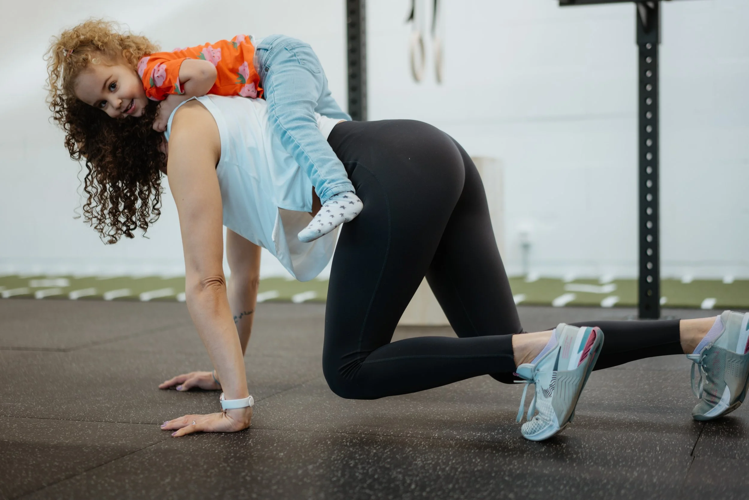 Eine Frau macht Übungen auf einer Gymnastikmatte, während ein kleines Mädchen auf ihrem Rücken liegt und sie anlächelnd ansieht.