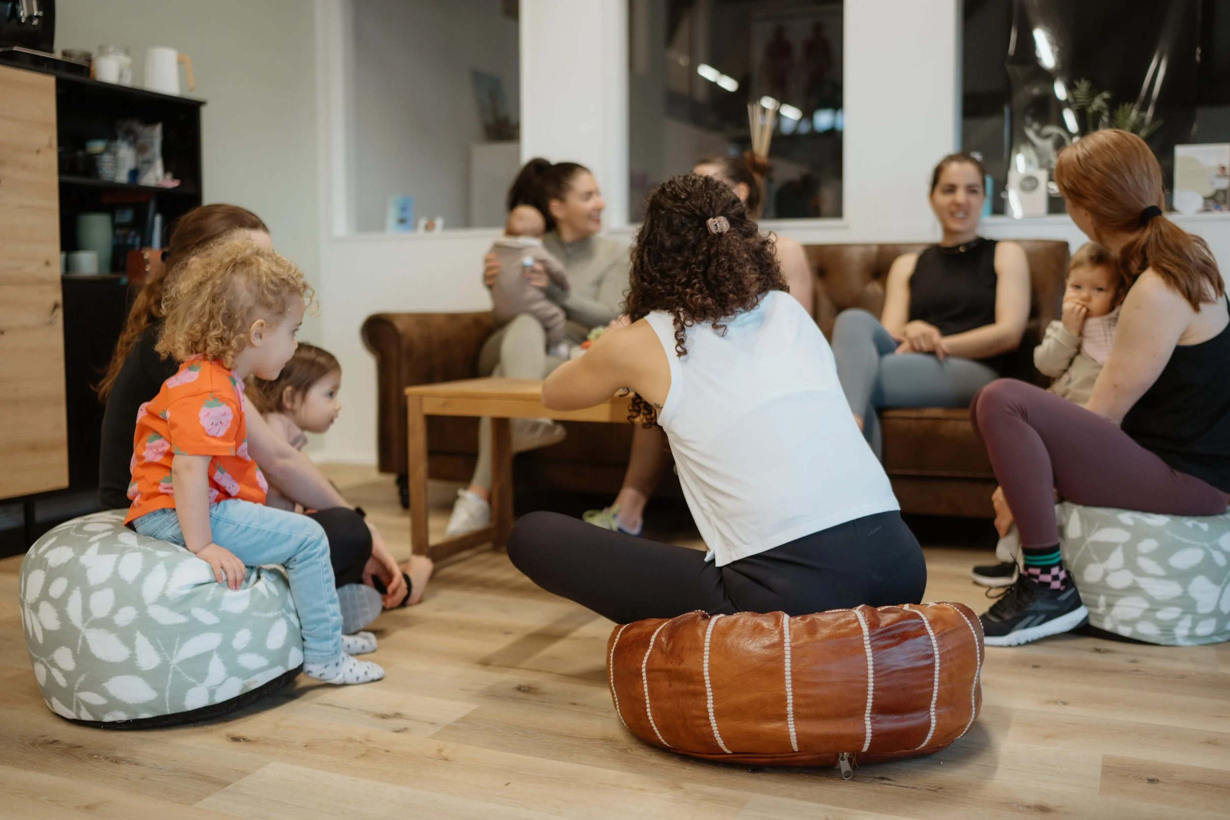 Mehrere Frauen und Kinder sitzen in einem Wohnzimmer, einige auf Hockern oder auf dem Boden, und unterhalten sich.