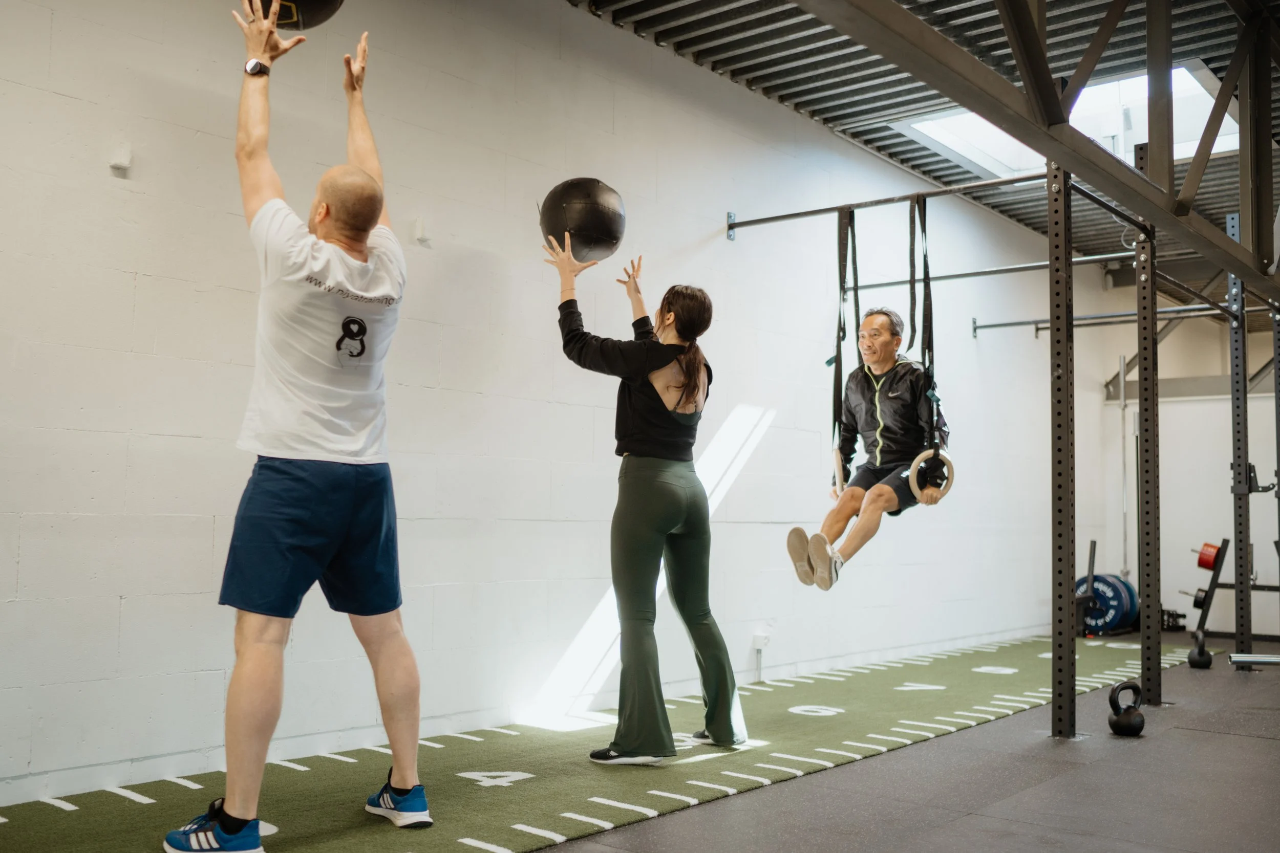 Drei Menschen trainieren im Fitnessstudio mit verschiedenen Übungen: einer springt mit Gymnastikringen, eine Frau hält ein Medizinball, ein Mann schwingt auf Turnringen.