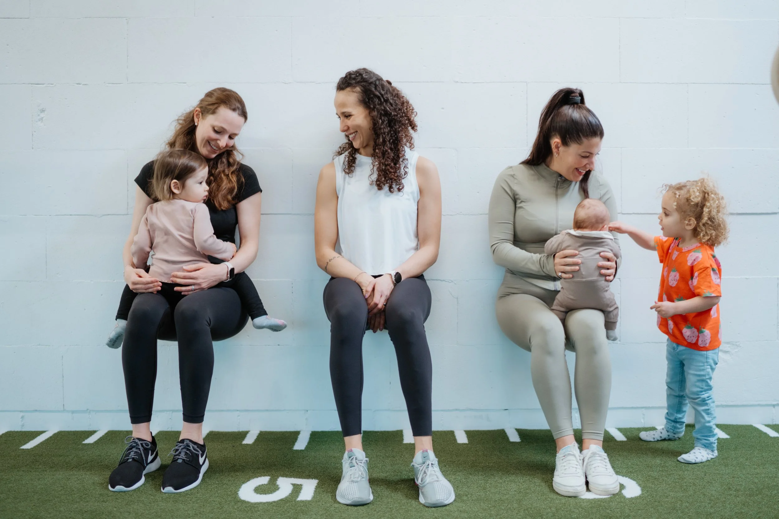 Drei Frauen sitzen an einer weißen Wand, begleiten ihre Kinder. Zwei Kinder spielen mit den Frauen, eines sitzt auf dem Schoß der ersten Frau, das andere wird von der dritten Frau gehalten, während die zweite Frau lächelnd zuschaut.