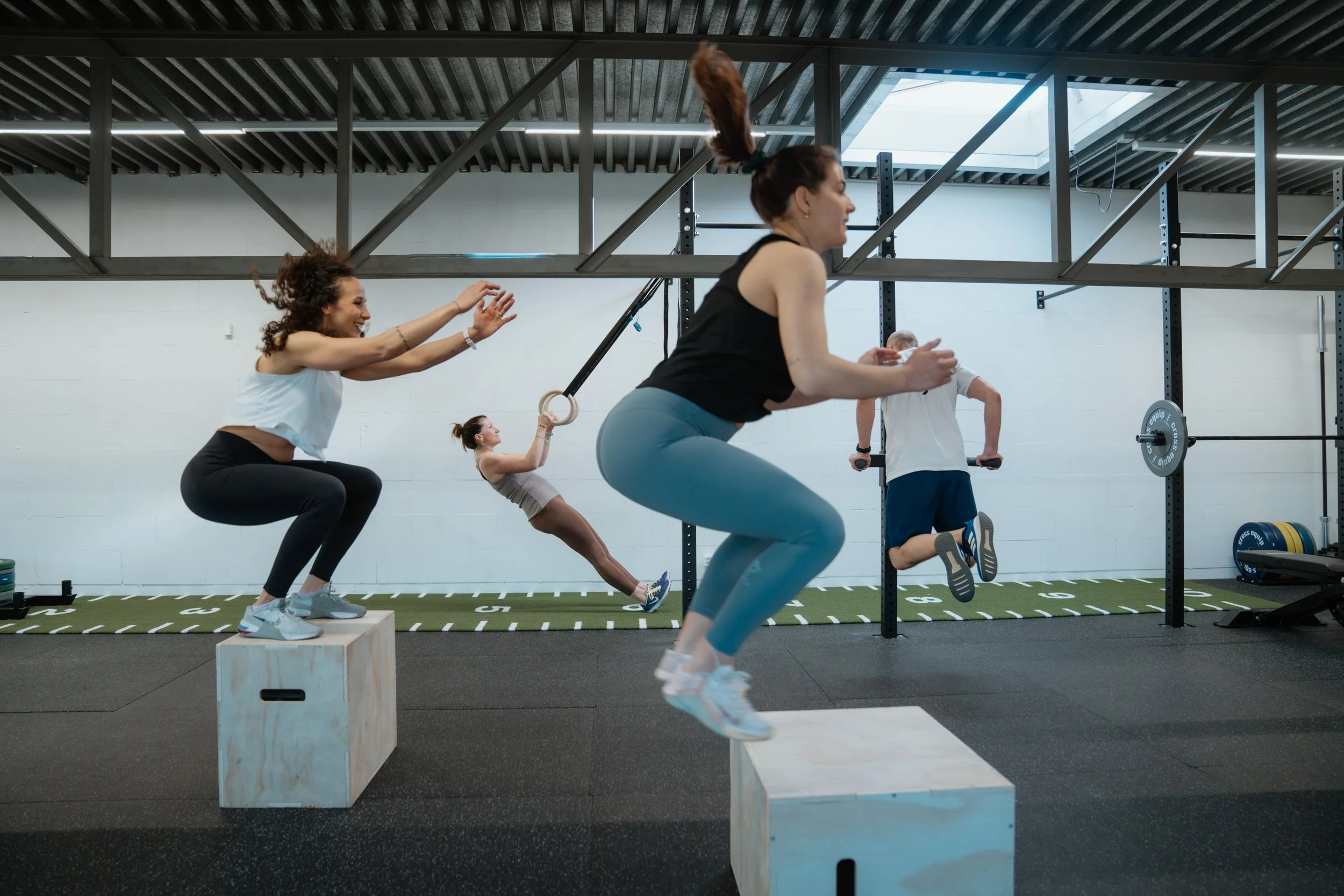 Menschen beim Workout in einem Fitnessstudio, darunter eine Frau springt auf eine Kiste, andere trainieren mit einem Springseil, Klimmzügen und Übungen mit Gewichten.