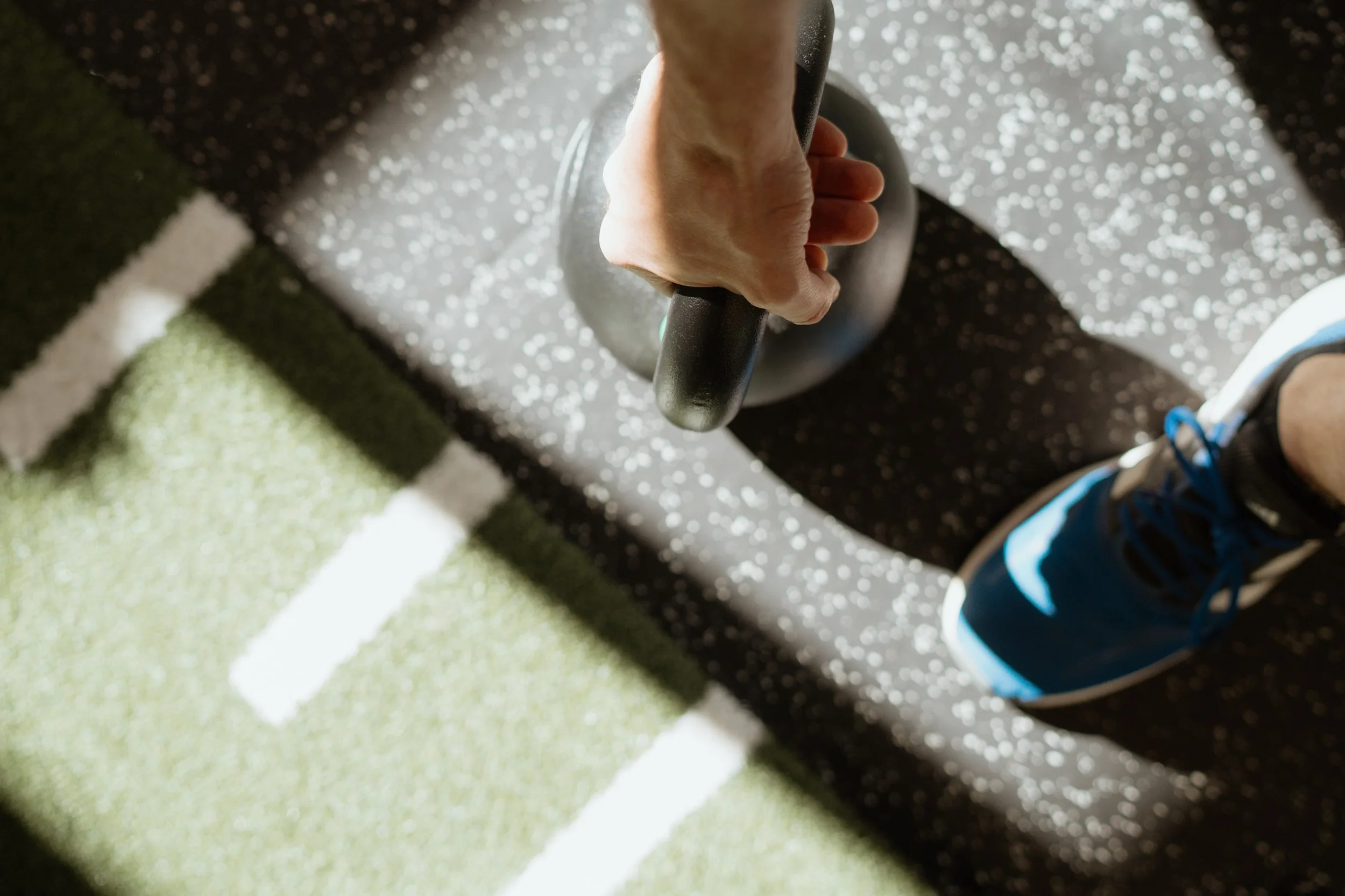 Person hebt eine Hantel im Fitnessstudio, Blick von oben, mit blauen Turnschuhen, auf einem schwarzen Trainingsboden mit weißen Sprenkeln und grüner Sportmatte