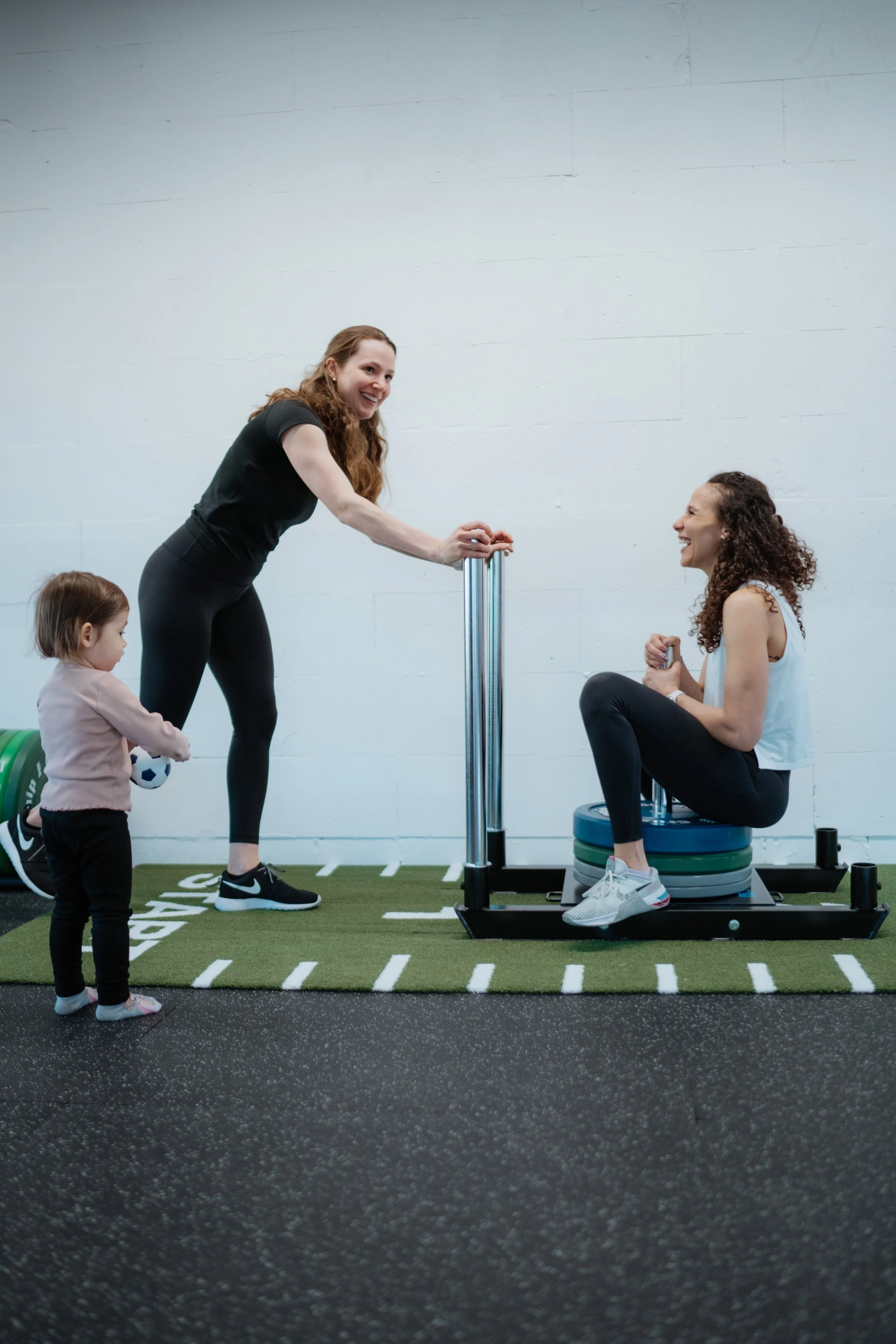 Zwei Frauen und ein kleines Mädchen in einem Fitnessstudio. Die eine Frau steht und hält eine Fitnessmaschine, während die andere auf einer Bank sitzt und lacht. Das kleine Mädchen steht in der Nähe und hält einen kleinen Ball.