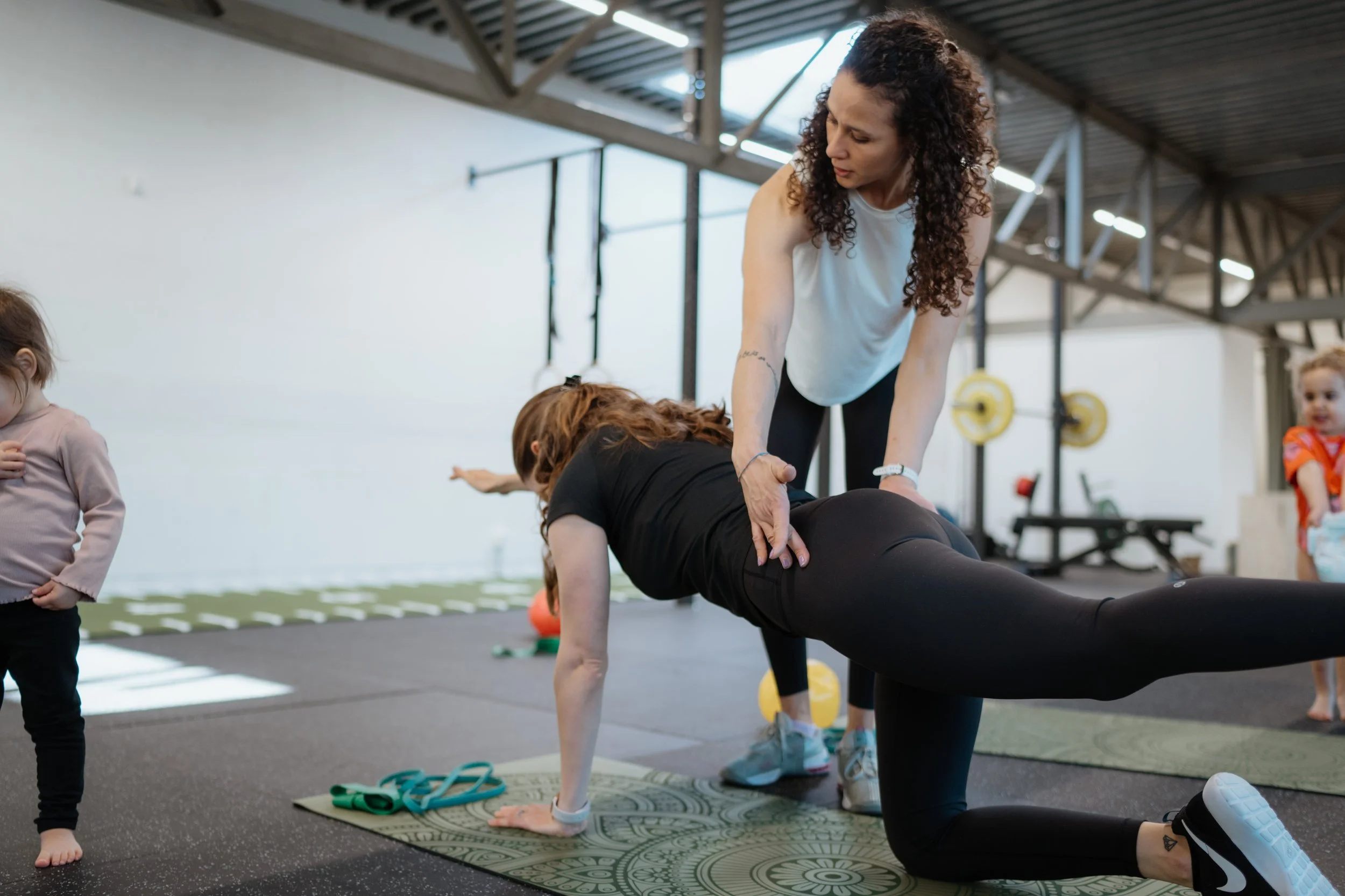 Eine Frau macht in einem Fitnessstudio mit Kindern Yoga, sie hilft einem Mädchen in eine Planke-Position, während andere Kinder im Hintergrund spielen.