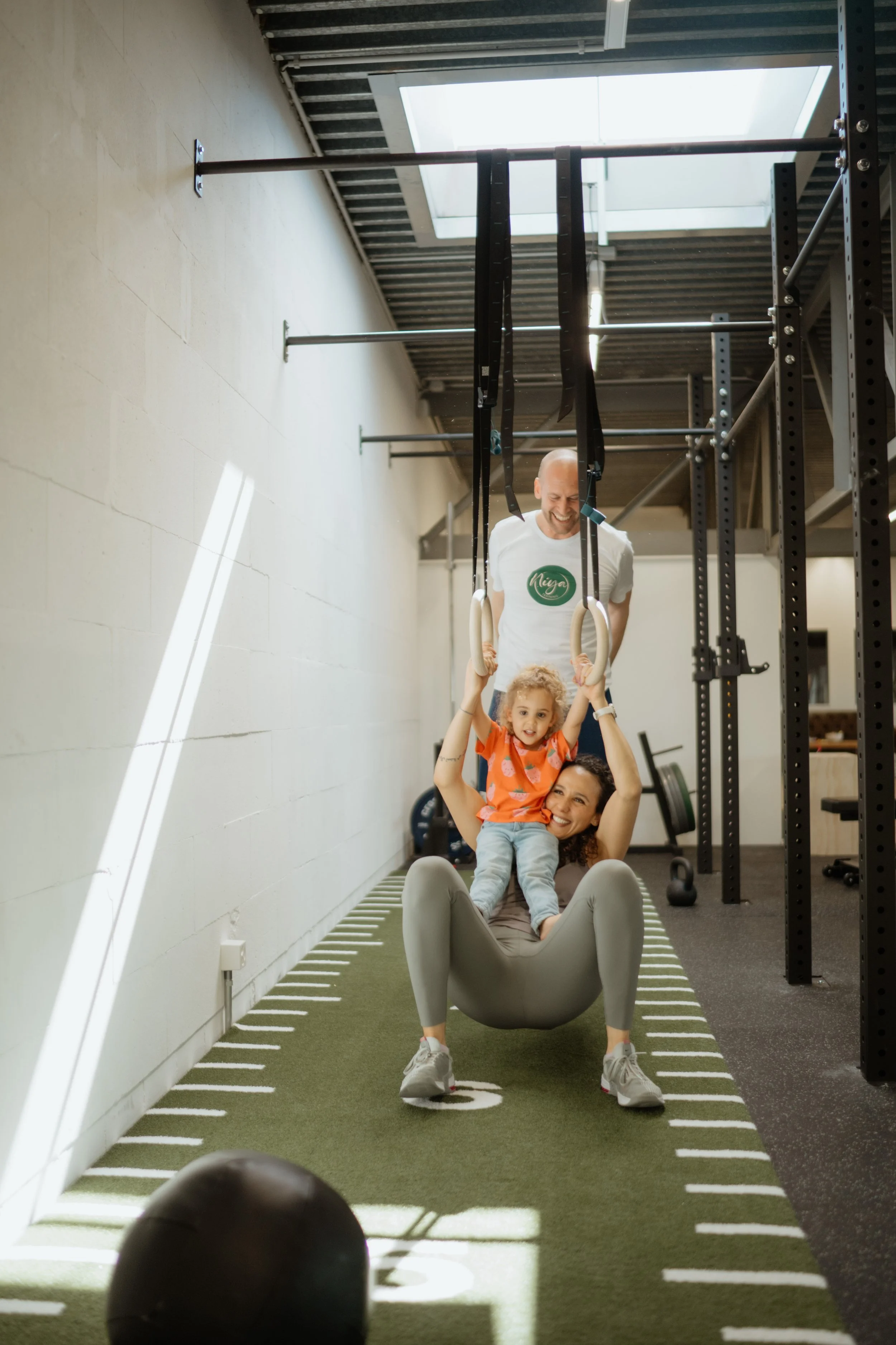 Familie beim Training in einem Fitnessstudio, die Mutter sitzt in einer Gymnastikhängematte, während das Kind an den Griffen der Ringe hängt, vom Vater unterstützt.