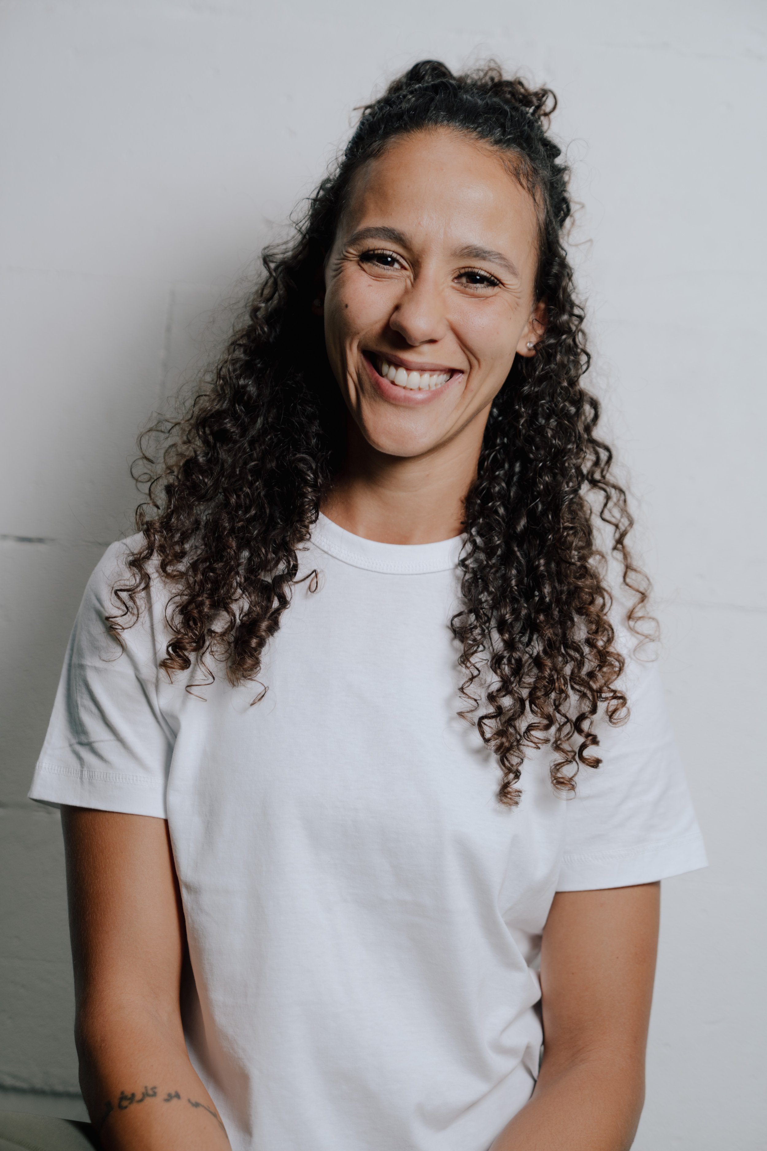 Lächelnde junge Frau mit lockigen Haaren in weißem T-Shirt vor weißer Wand.