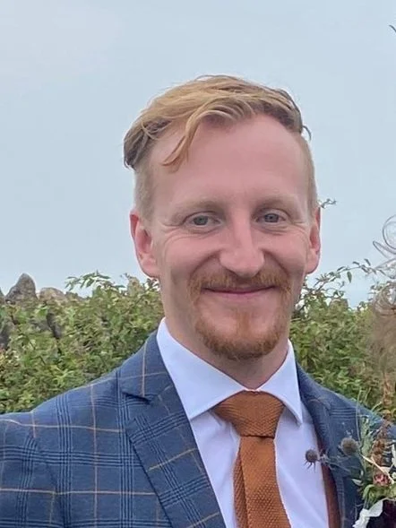 A smiling man with light hair and a beard, dressed in a blue plaid suit, white shirt, and brown tie, outdoors with greenery in the background.