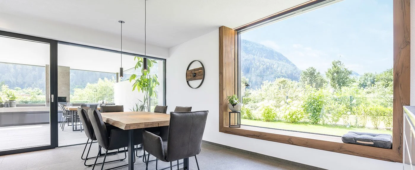 Dining room with a large wooden table, black chairs, and a floor-to-ceiling window showing a lush green landscape and mountains outside.