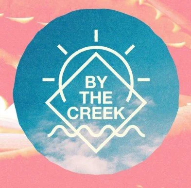 A circular logo with a blue background featuring a white geometric mountain, sun, and wave illustrations, with the text "BY THE CREEK" in the center.