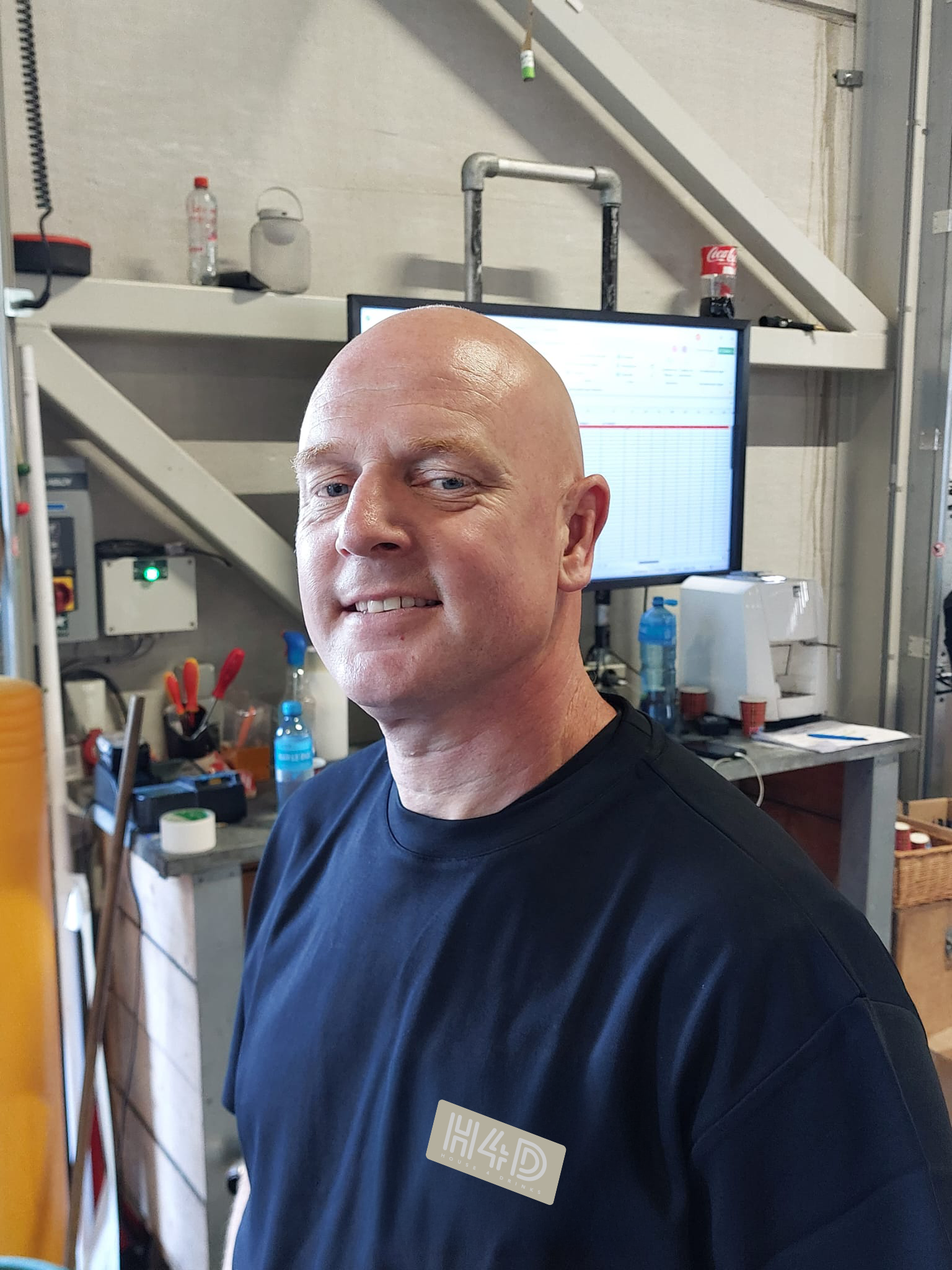 A bald man smiling in a workshop or industrial setting, with a computer monitor and various tools in the background.